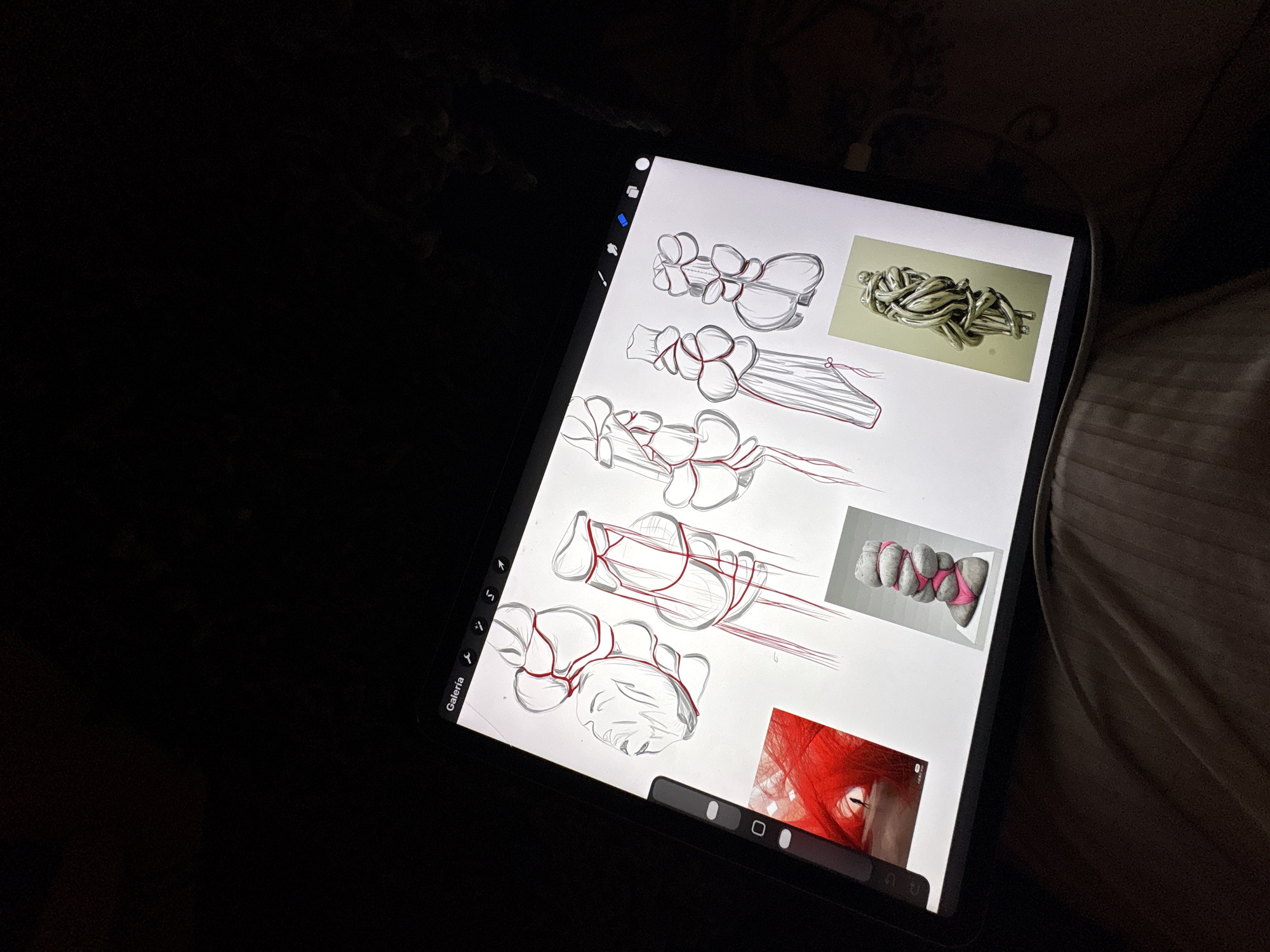 The image displays an illuminated digital tablet screen in a dimly lit environment, suggesting a nighttime or low-light setting. The tablet, likely an iPad Pro, is seen from a slightly elevated angle, resting on a surface partially covered by creased, light brown fabric on the right side of the frame, possibly a bed or sofa.
The tablet screen showcases a digital workspace filled with design sketches and reference images. The main subjects on the digital canvas are multiple conceptual drawings, rendered in grey lines with distinctive red overlays or structural accents. These sketches depict various iterations of abstract, organic, or interconnected pod-like forms, hinting at designs for jewelry, accessories, or sculptural pieces.
Three smaller reference images are embedded within the digital canvas. In the top-right, there is a photograph of tangled, silver-colored metallic strands or wires, resembling a complex knot or abstract sculpture. Below this, a second image displays several grey, elongated, pebble-like objects bound together with bright pink straps or fabric. The third reference image, located in the bottom-right, is a close-up of something intensely red and textured, possibly fur or textile, featuring a pale, almond-shaped eye.
The tablet's interface shows a vertical toolbar with various digital art tools on the left side, and a horizontal bar at the bottom center, likely for brush or color selection. The text "Galería" is visible at the bottom-left corner of the screen, indicating a gallery function.
The scene captures a moment of creative activity, where an individual is engaged in digital design, possibly developing concepts for fashion, product design, or art, a fitting endeavor given the context of Milan, Italy.