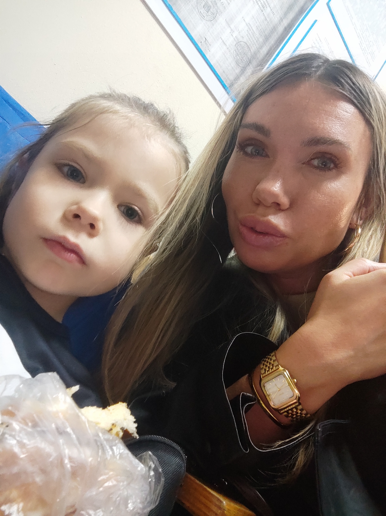 Two individuals, a woman and a young child, are positioned close together. The child is to the left and slightly in front of the woman, and both are facing the camera. The child has fair skin, light-colored hair, and a neutral expression. The woman, on the right, has tan skin, long blonde hair, and wears a gold watch and hoop earrings. They appear to be inside a room with wall-mounted documents visible in the background. The child holds a pastry or bread wrapped in plastic in their lap. The presence of wall-mounted documents suggests the setting could be an office or waiting area, but without more information, the specific location in Votkinsk, Russia cannot be determined.