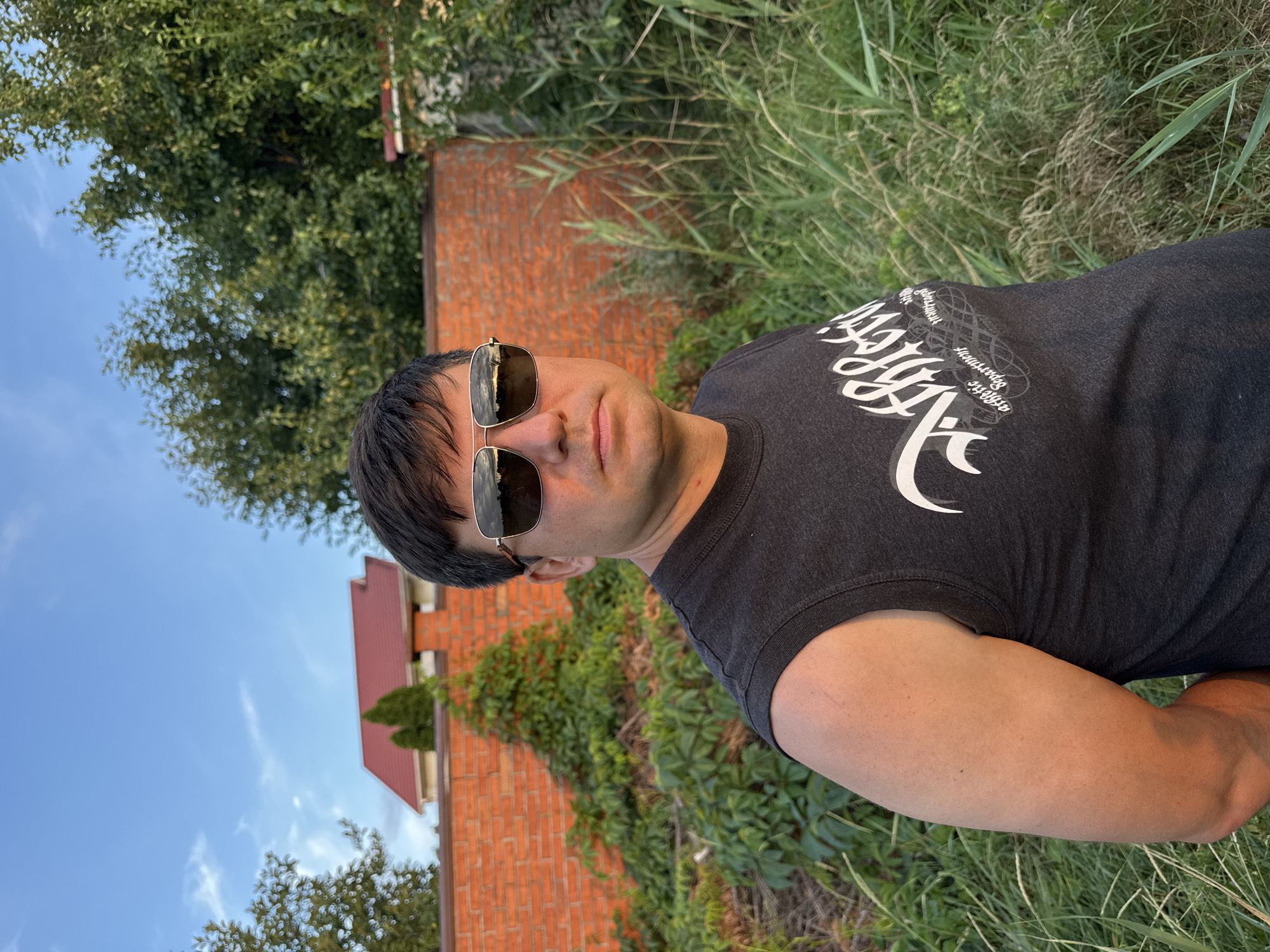A man wearing a dark grey sleeveless t-shirt and aviator sunglasses stands outdoors, facing slightly to the right. He has short, dark hair and fair skin. The t-shirt has white graphic text on it. Behind him, a tall brick wall runs horizontally, with lush green vegetation growing against it and in front of it. To the left, a red-roofed building is partially visible above the wall. The sky is blue with scattered white clouds. The ground is covered in tall green grass. The scene appears to be a residential backyard or a similar outdoor space in the village of Karachunivka, Ukraine.