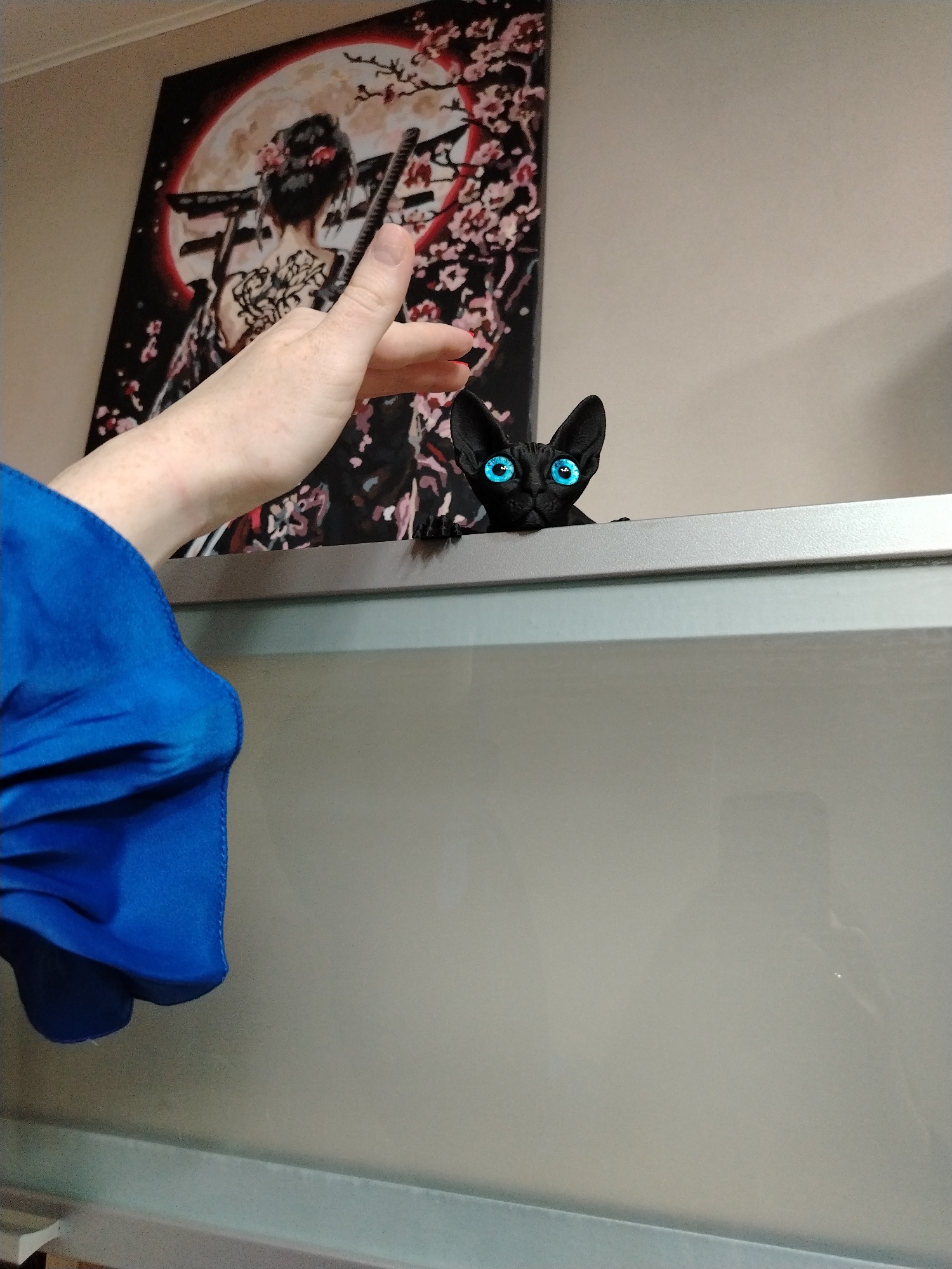 A medium shot captures a scene within an interior setting. A blue-eyed black cat is visible, with only its head and paws extending above the edge of a gray rectangular structure. To the left of the cat, a person's hand and part of a blue sleeve are visible. Behind the cat and hand, a poster featuring an image resembling a Japanese geisha with a sword is affixed to the wall. The background wall is a light beige color. The location is likely indoors, but the city of Izhevsk, Russia cannot be confirmed from this image alone. There are no apparent actions or interactions taking place.