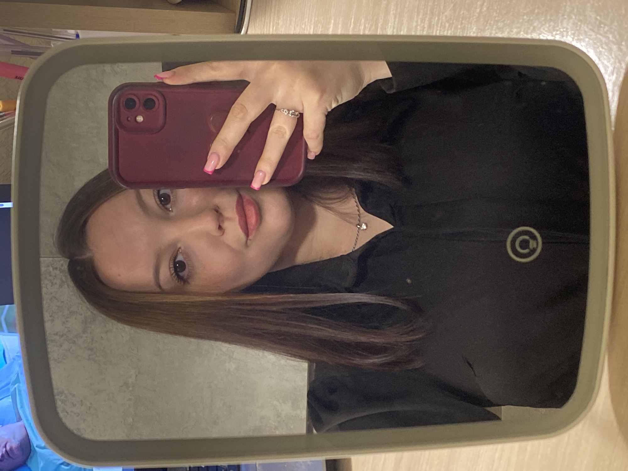 A person takes a mirror selfie with a maroon-colored iPhone 11. The person has shoulder-length brown hair, light skin, and wears a black top with a round logo on the right side. They are wearing a ring on their left hand. The mirror has a light green frame. The background shows a glimpse of a computer monitor with a blue image, and part of a wooden shelf. The location is likely indoors, but there is no indication of Chaikovsky, Russia.