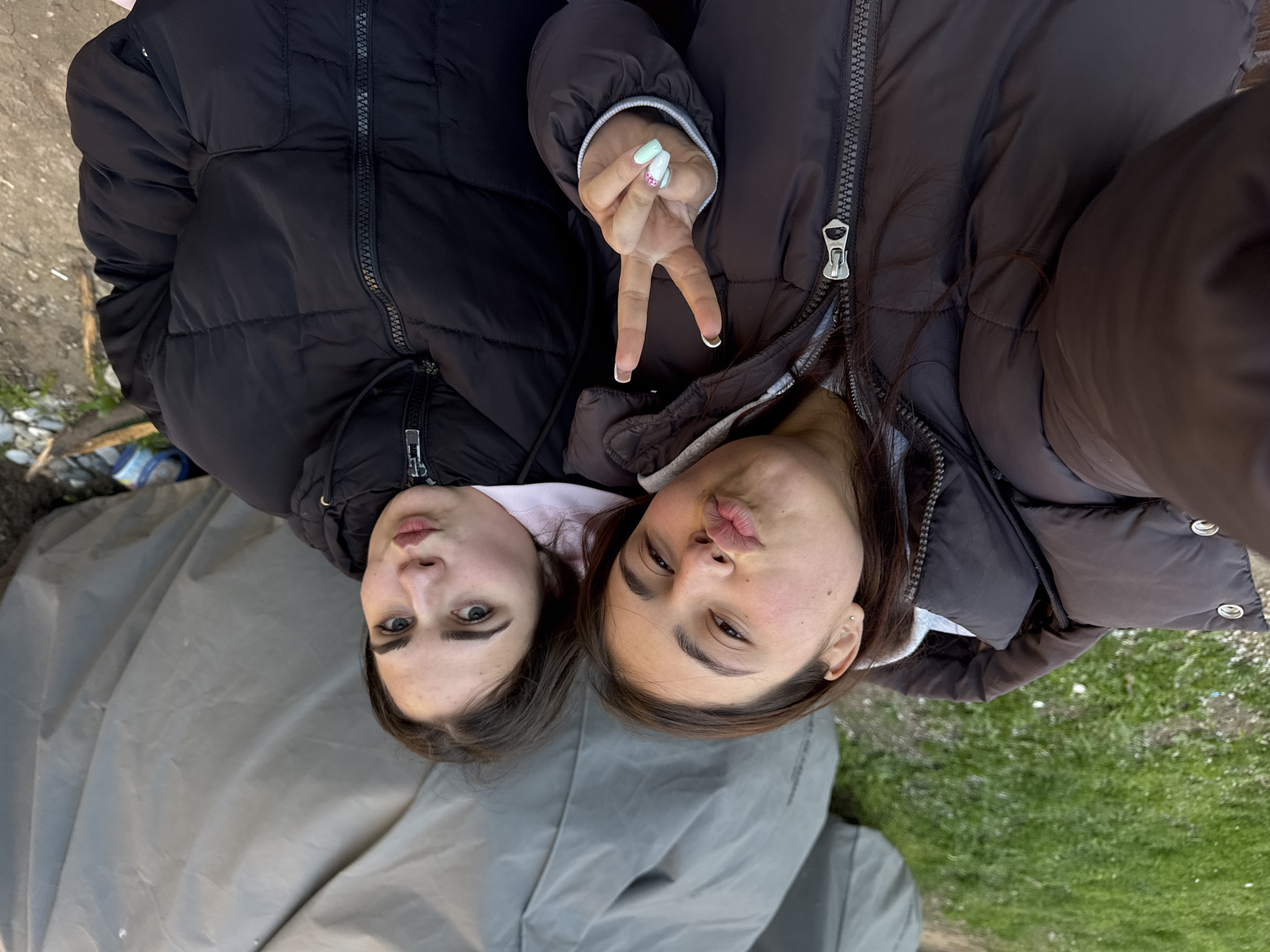Two young women are posing for a selfie, lying on what appears to be a grey tarpaulin. The woman on the left has dark brown hair, fair skin, and is wearing a black puffer jacket. The woman on the right has dark brown hair, light brown skin, and is wearing a brown puffer jacket, holding up two fingers in a peace sign. Both are pouting their lips toward the camera. The location is outdoors, with greenery visible in the bottom right corner, suggesting grass. Part of a concrete or asphalt surface is visible in the upper left. The image does not provide enough information to determine the specific location within the city of Sofia, Bulgaria.