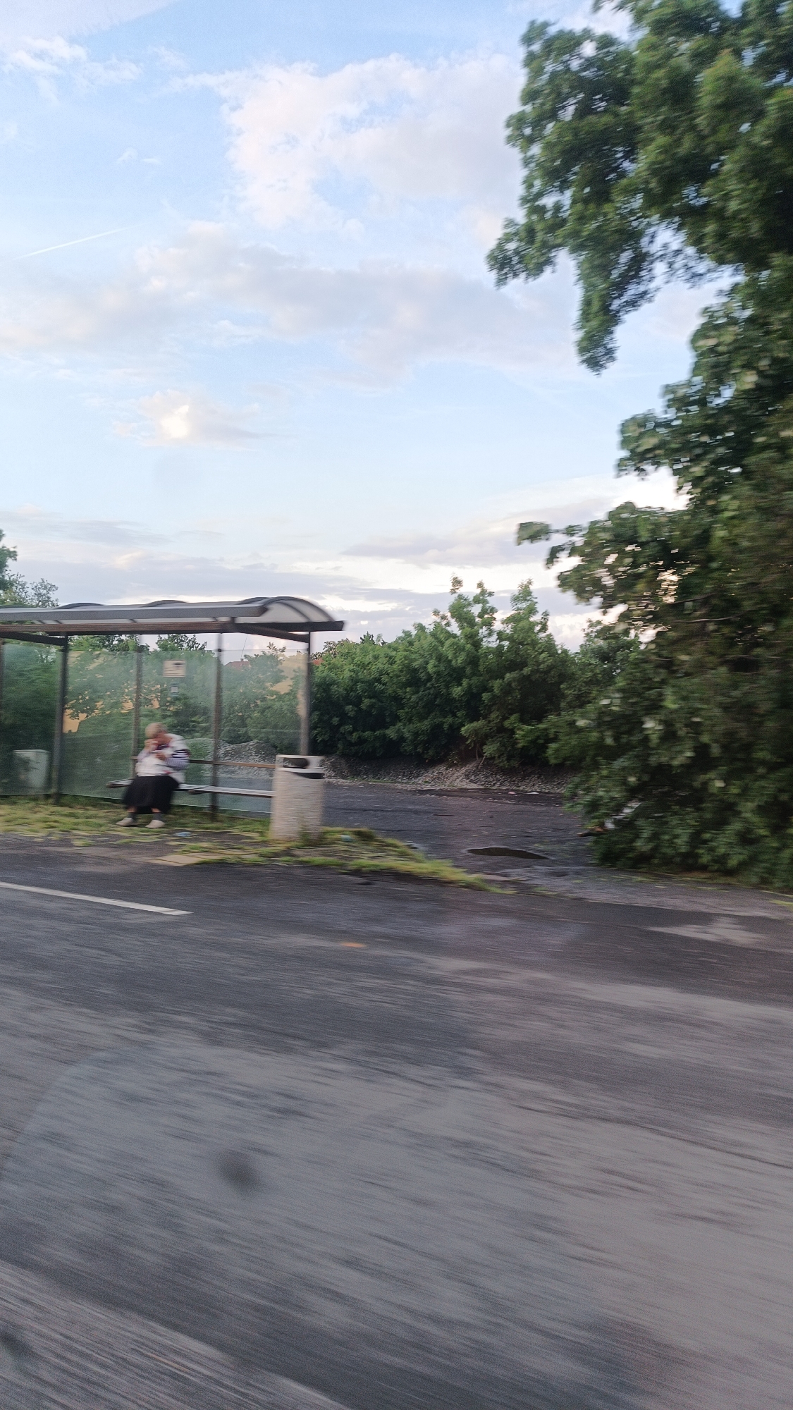 The image depicts an outdoor scene likely captured from a moving vehicle. A bus stop shelter is visible on the side of a dark, asphalt road. A woman is seated inside the shelter. A grey trash can stands adjacent to the shelter. Behind the bus stop, there's dense green vegetation and trees. The sky is partly cloudy with a pale blue hue. The blurring suggests the image was taken while in motion. It is not possible to determine the precise location within Burgas, Bulgaria, from the visual information provided.