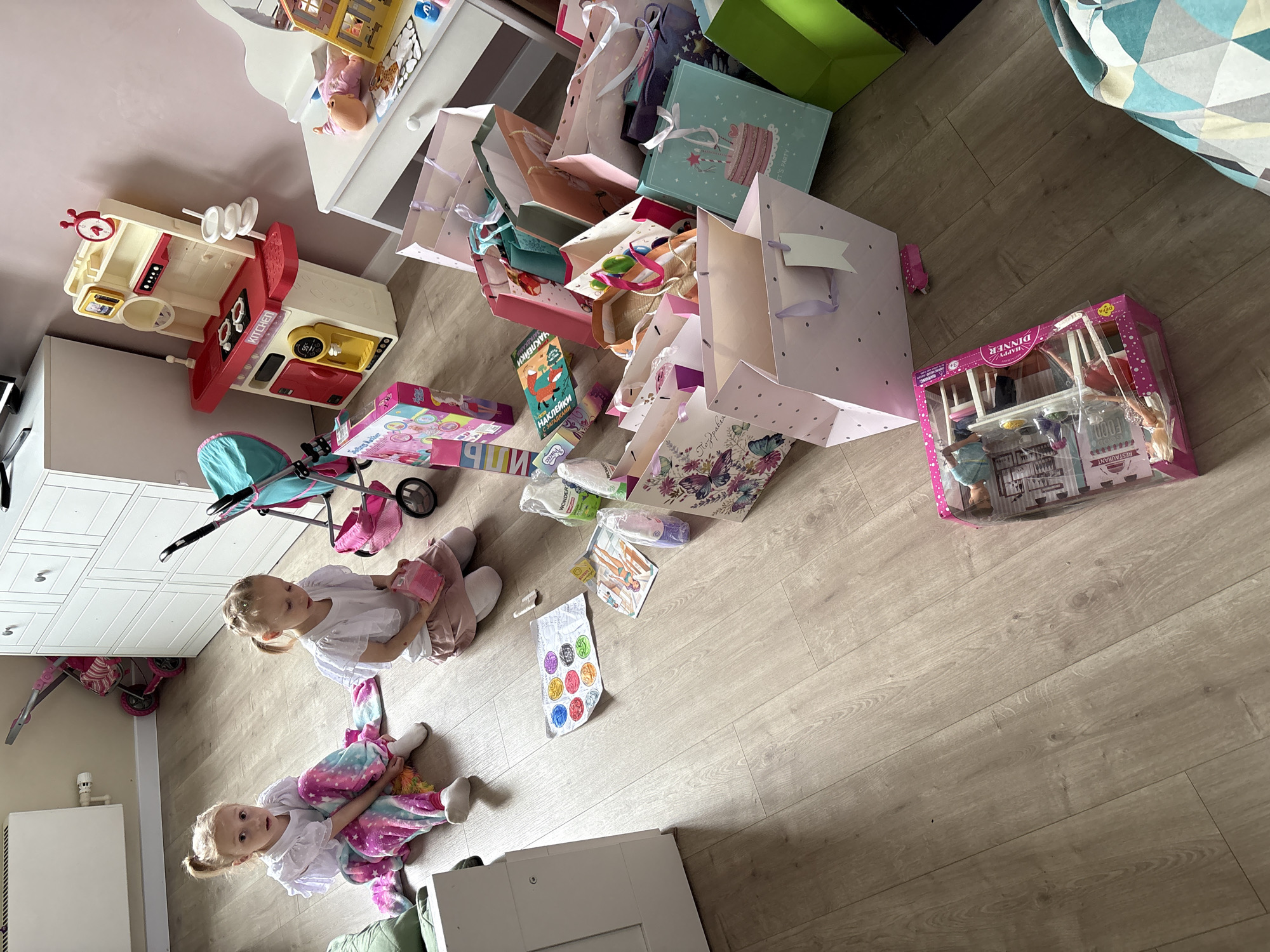 The image depicts an indoor scene, possibly a child's room, strewn with toys and gift bags. Two young girls are sitting on the floor amid the clutter. One girl is wearing a white top and multicolored pants, while the other is wearing a white top and pink pants. Numerous gift bags are scattered around the room, along with various toys, including a toy kitchen set, a doll stroller, a dollhouse, and a sticker sheet. The room appears to be located on the territory of Krasnoyarsk, Russia because some of the text on the toys is in Russian. The floor is made of light-colored wood.