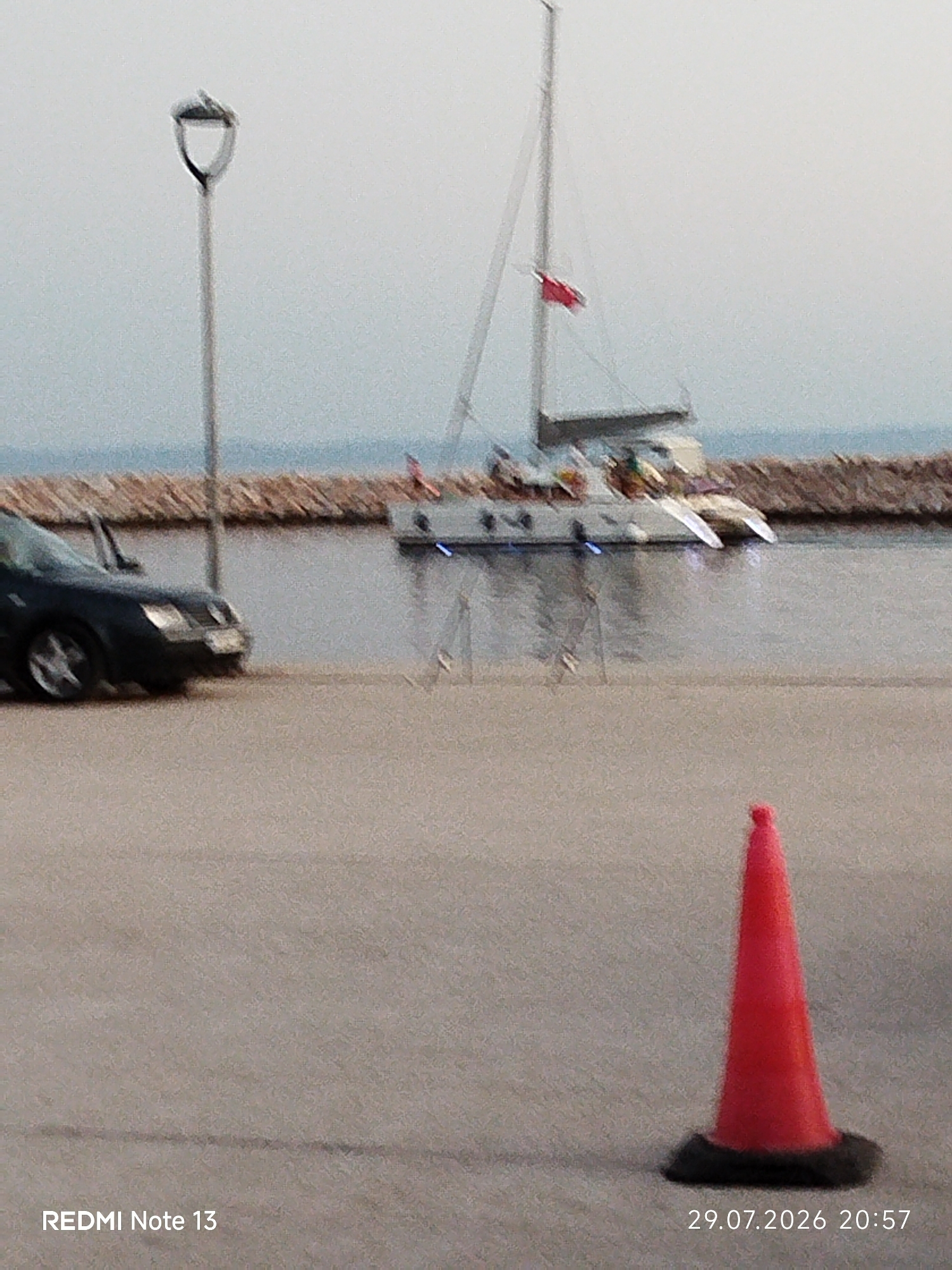 A red traffic cone is in the foreground, casting a shadow on the pavement. In the background, a dark car is parked next to a body of water. A catamaran is anchored in the water, and a stone breakwater runs along the shoreline. A lamppost stands to the left of the frame, and the sky is overcast. The image was taken in Sithonia Municipal Unit, Greece.