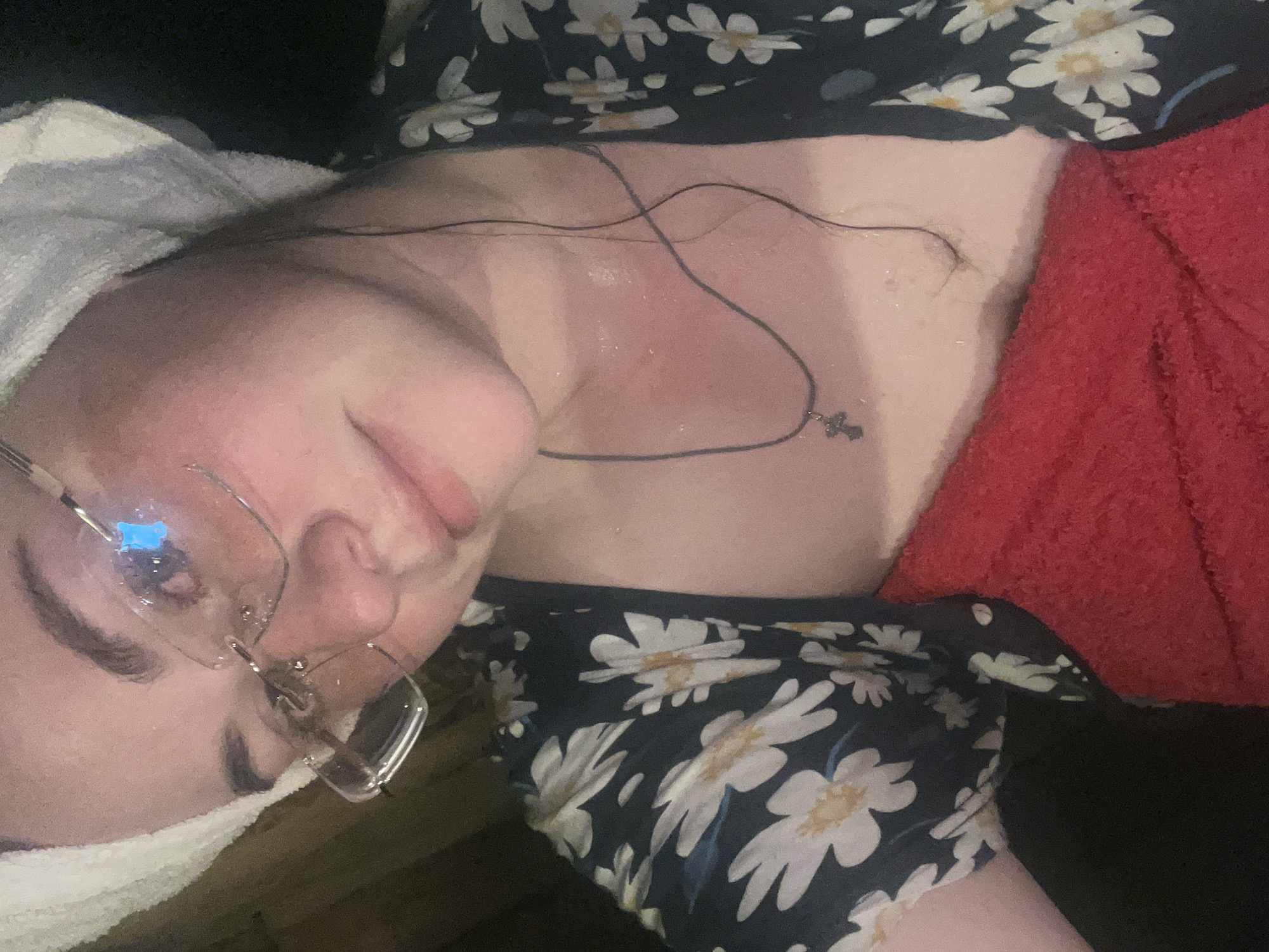 A woman lies at an angle, wearing glasses, a towel wrapped around her head, a daisy-patterned top, and a necklace with a cross pendant. A red towel is visible on her right side. The location of Сосново, Russia, cannot be determined from the visual information.