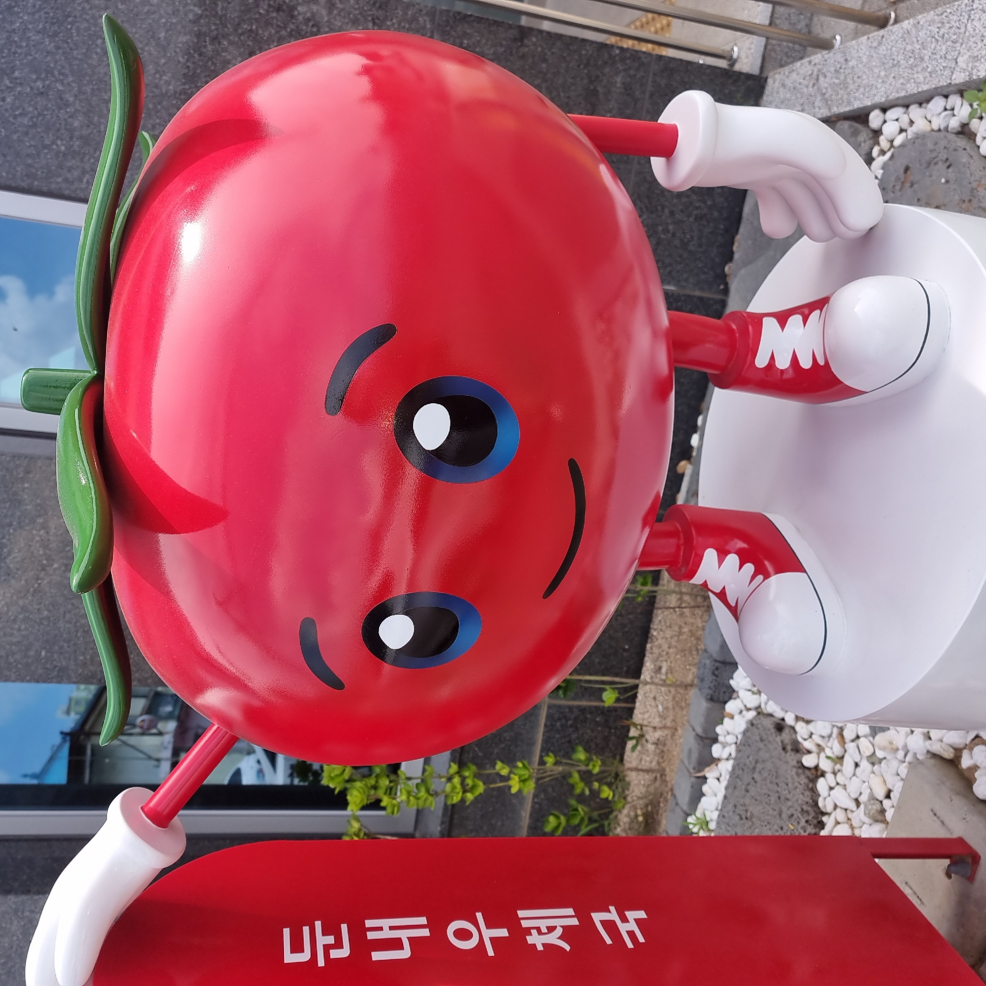 A cartoonish, oversized tomato sculpture stands on a white cylindrical pedestal in front of a building with a dark, textured facade. The tomato is bright red with a green stem and leaves, and features a cheerful face with large blue and white eyes and a smiling mouth. It has red stick-like legs and feet shaped like white sneakers with red laces. The sculpture is positioned to the right of a tall, red, rectangular object with white Korean text that reads "눈 내 우체국" (Nunnae Post Office). The ground around the pedestal is covered with white pebbles and some darker stones. Green foliage is visible behind the sculpture. The environment appears to be outdoors, suggesting a public space such as a post office or a town square in Dunnae-myeon, South Korea.