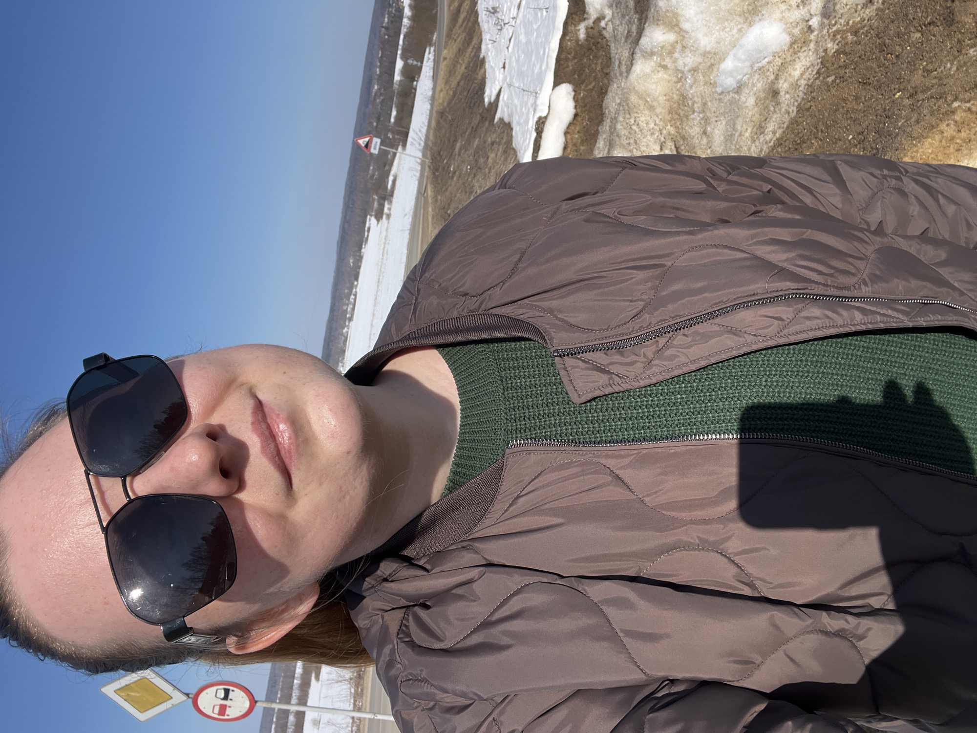 A female subject is depicted in an outdoor setting in Zubovo, Russia. She is shown from the chest up, wearing dark aviator-style sunglasses, a dark green knitted sweater, and a brown quilted bomber jacket with a distinct quilted pattern and ribbed collar. Her hair is pulled back, and her facial expression is neutral with a slight upturn at the corners of her mouth.
To the left of the subject, behind her left shoulder, two Russian road signs are visible on a single pole: an upper yellow diamond sign indicating a 'priority road' and a lower circular red-bordered sign with two cars crossed out, signifying 'no overtaking.' The background features a snow-covered landscape with visible patches of exposed brown earth, indicating melting conditions. A clear blue sky dominates the upper portion of the frame. In the mid-ground to the right, a red triangular warning sign with a white background and a '300m' notation is partially visible, likely indicating a road hazard such as a hill. Distant trees and what appears to be a small settlement are discernible on the horizon. The immediate foreground in the lower right consists of dirty, melting snow and brown earth. The scene is brightly illuminated by direct sunlight, casting a shadow consistent with a handheld device onto the subject's jacket in the lower left.