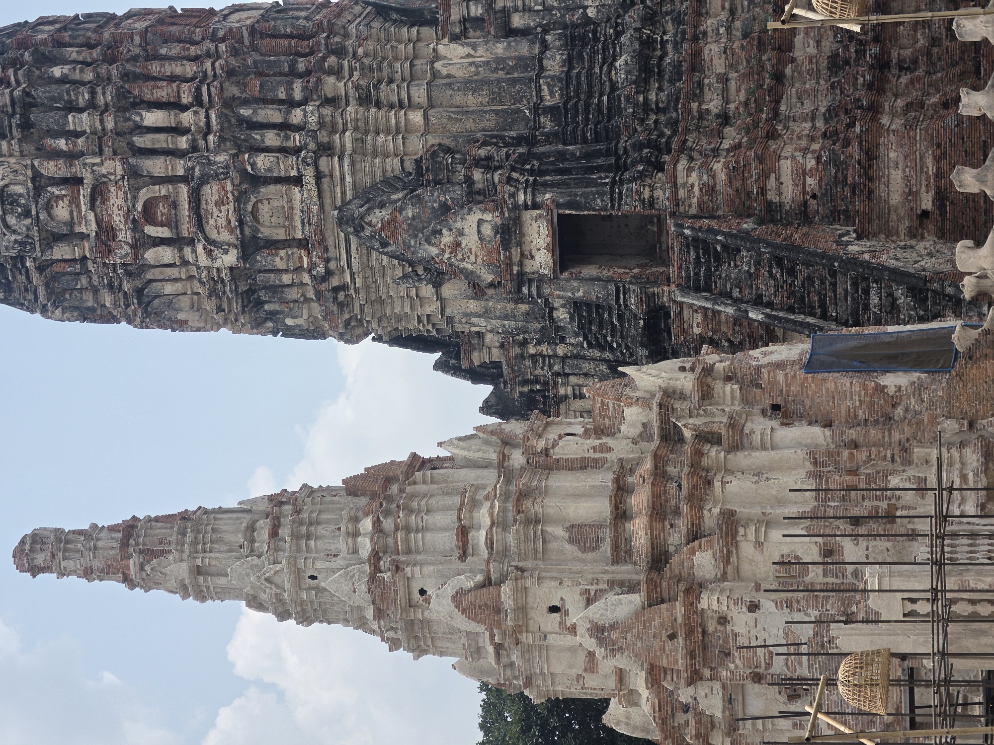The image shows Wat Yai Chaimongkol, located in Ban Pom Subdistrict, Thailand, featuring the crumbling, brick structures of a historical temple complex, with two prominent spires rising against a backdrop of a cloudy sky. One spire is in a state of deterioration, revealing layers of brick, while the other is darker and more weathered. Restoration efforts appear evident, as scaffolding and woven baskets are visible on the structure.