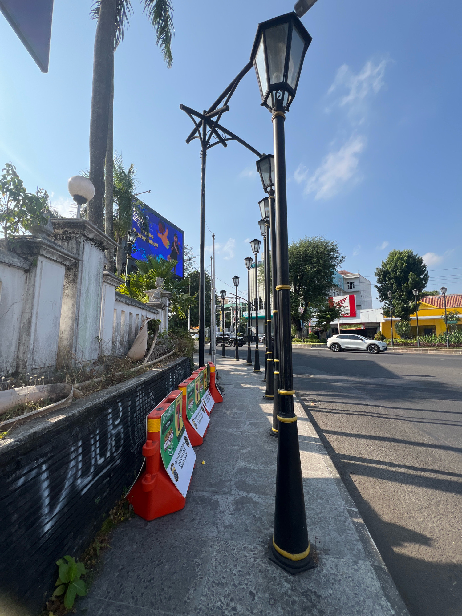 This is a street-level, eye-level, medium-wide shot of a sidewalk and road on a sunny day in the city of Yogyakarta, Indonesia. The sidewalk is made of concrete pavers and runs along a black, graffiti-covered low wall on the left. A row of orange traffic barriers with white signs is placed along the edge of the sidewalk. Several black lamp posts with ornate bases and gold accents line the sidewalk, casting shadows on the ground. Beyond the sidewalk, a paved road is visible with a white car driving on it and a yellow building with an orange roof in the background. In the distance, trees, another building with a red and white Honda sign, and a large, blue digital billboard are visible under a clear blue sky with sparse clouds. A palm tree grows near the wall on the left, and dry leaves and debris are present on the ground.