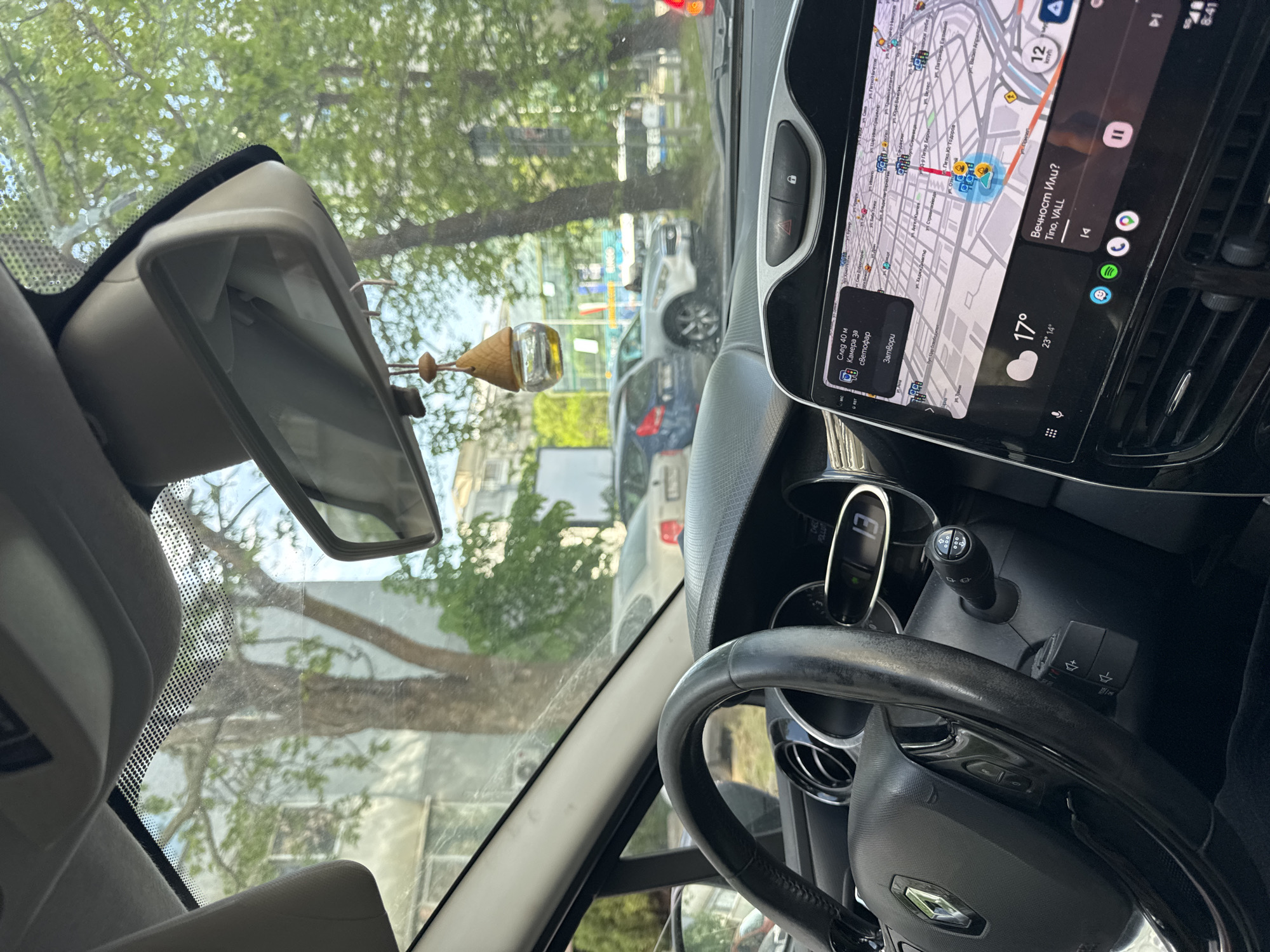 A view from inside a car shows the dashboard with a navigation system displaying a map of Varna, Bulgaria. The map includes "Вечност Или?" and the location of "Tino, VALL". The weather is displayed as 17 degrees Celsius, ranging from 23 to 14 degrees. The navigation indicates a speed of 12 km/h, a camera for traffic lights in 40 meters. Street names visible on the map include "ул. Цар Освобод" (Tsar Osvoboditel street), "Д-р Петър Берон" (Dr. Petar Beron street), and "бул. Петко Ю. Тодоров" (Petko Y. Todorov Blvd). The rearview mirror and steering wheel are visible. Through the windshield, trees and parked cars on the street can be seen.