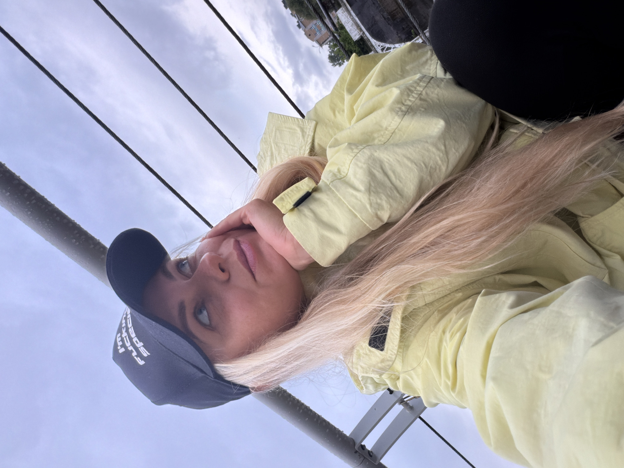 A young adult female, with long blonde hair falling over her right shoulder, is depicted in a close-up selfie taken outdoors in Votkinsk, Russia. She wears a black baseball cap with the white embroidered text "I'M FUCKIN' SPECIAL" and a light yellow windbreaker jacket. Her right hand supports her chin as she looks upwards and slightly to the right with a pensive expression. The background features a heavily overcast sky with grey clouds. Several dark, taut cables or ropes extend diagonally across the upper and middle portions of the frame, parallel to a thick, dark grey metallic railing visible on the left, which shows visible water droplets. In the far right background, trees and a light-colored multi-story building with a red roof are faintly visible through the cables, suggesting an urban or semi-urban environment. Her black leggings are visible in the lower right corner.
