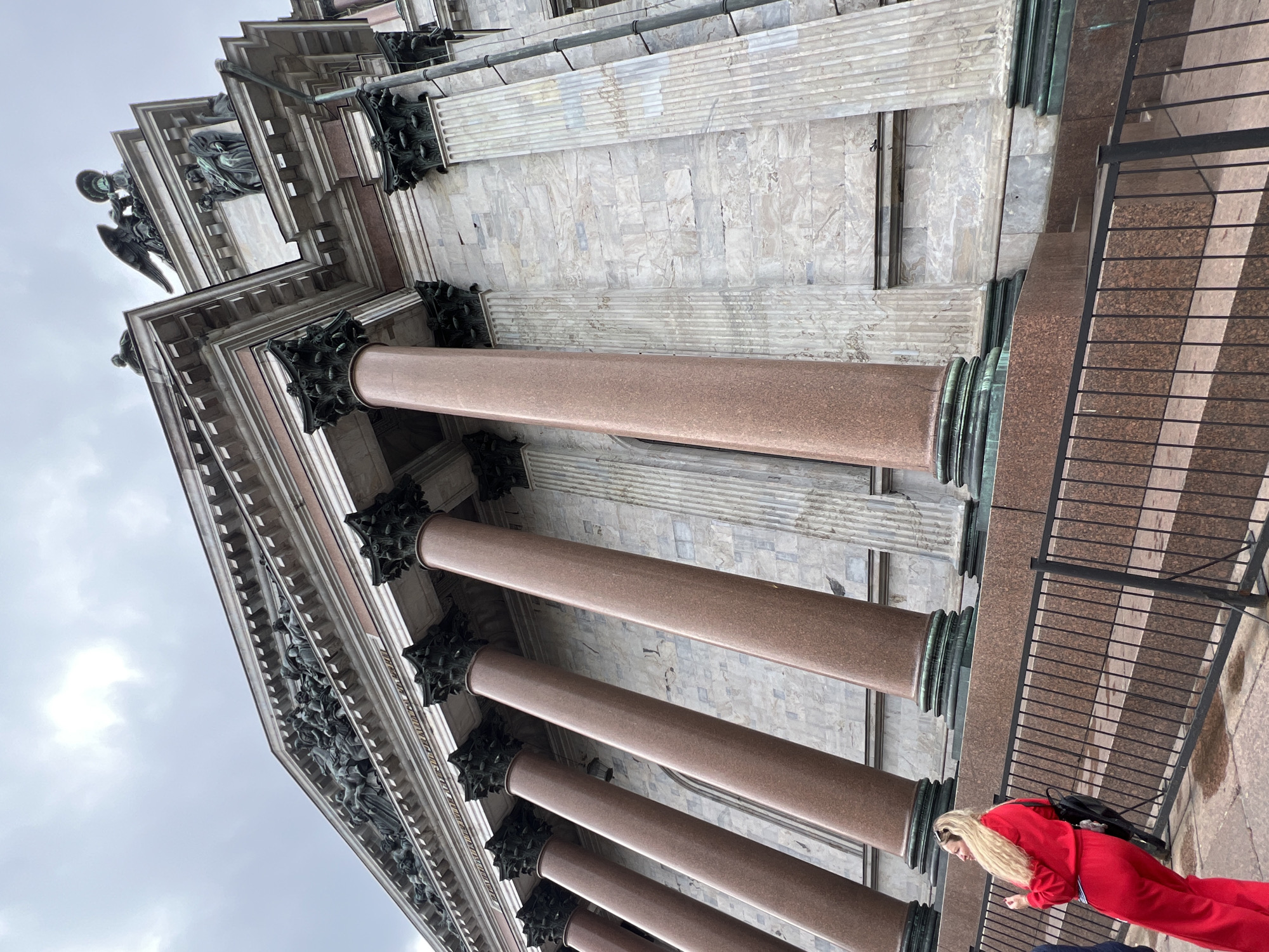 The image captures a low-angle perspective of the Saint Isaac's Cathedral in Saint Petersburg, Russia. The architecture showcases massive, polished red granite columns adorned with ornate Corinthian capitals made of bronze. Above the columns, a decorative frieze is visible, featuring sculptures. At the top of the structure, statues of angels.
A woman in a red pantsuit is standing near a metal railing. She has long blonde hair and is wearing a black backpack. The scene suggests a cloudy day.