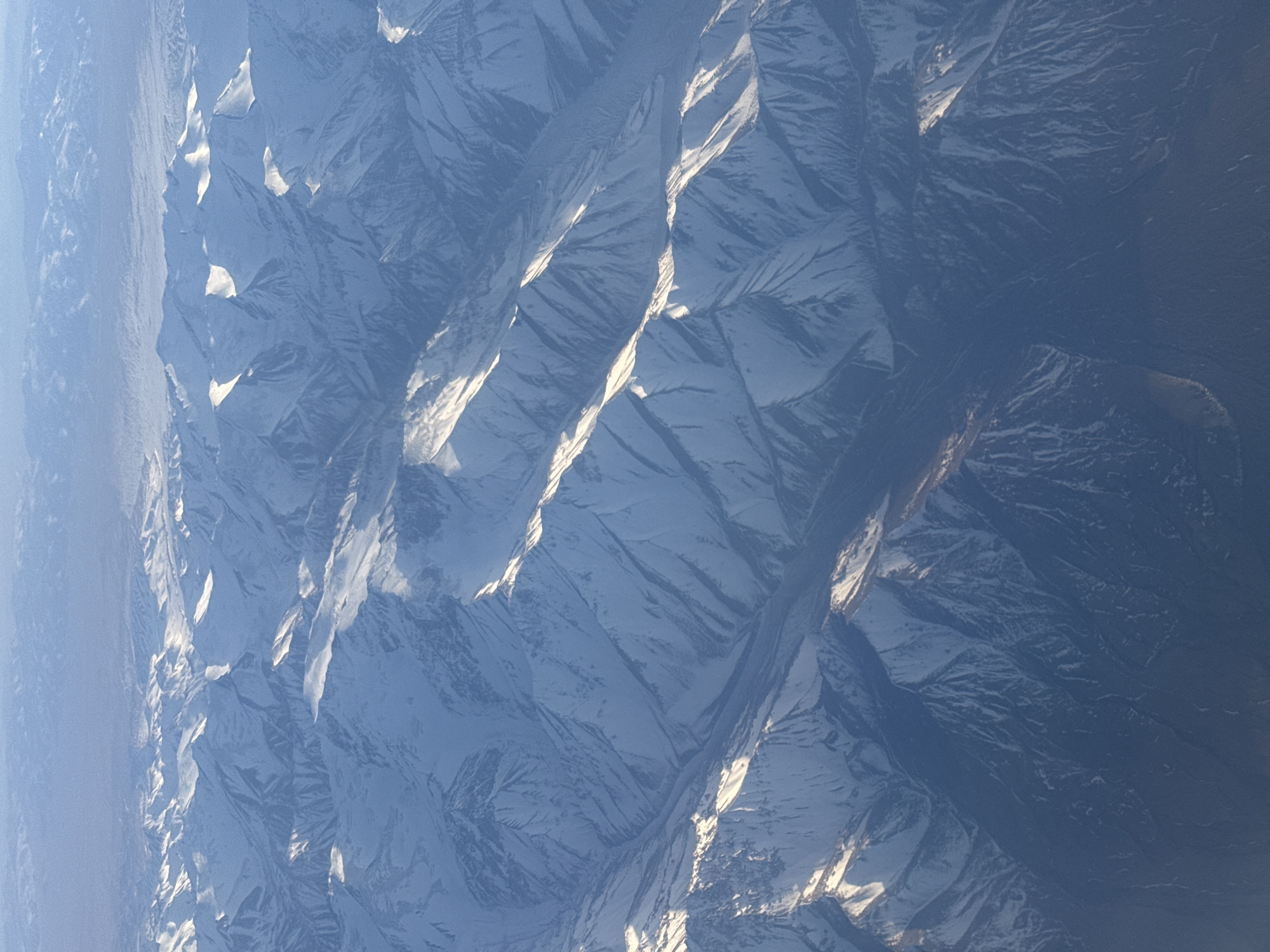 The image shows a mountainous region covered in snow. The mountain ridges are prominent, with shadows and sunlight creating contrasting patterns on the snow-covered surfaces. There are no visible signs of human presence or infrastructure. The location is unknown. The city of Tashkent, Uzbekistan is not depicted.