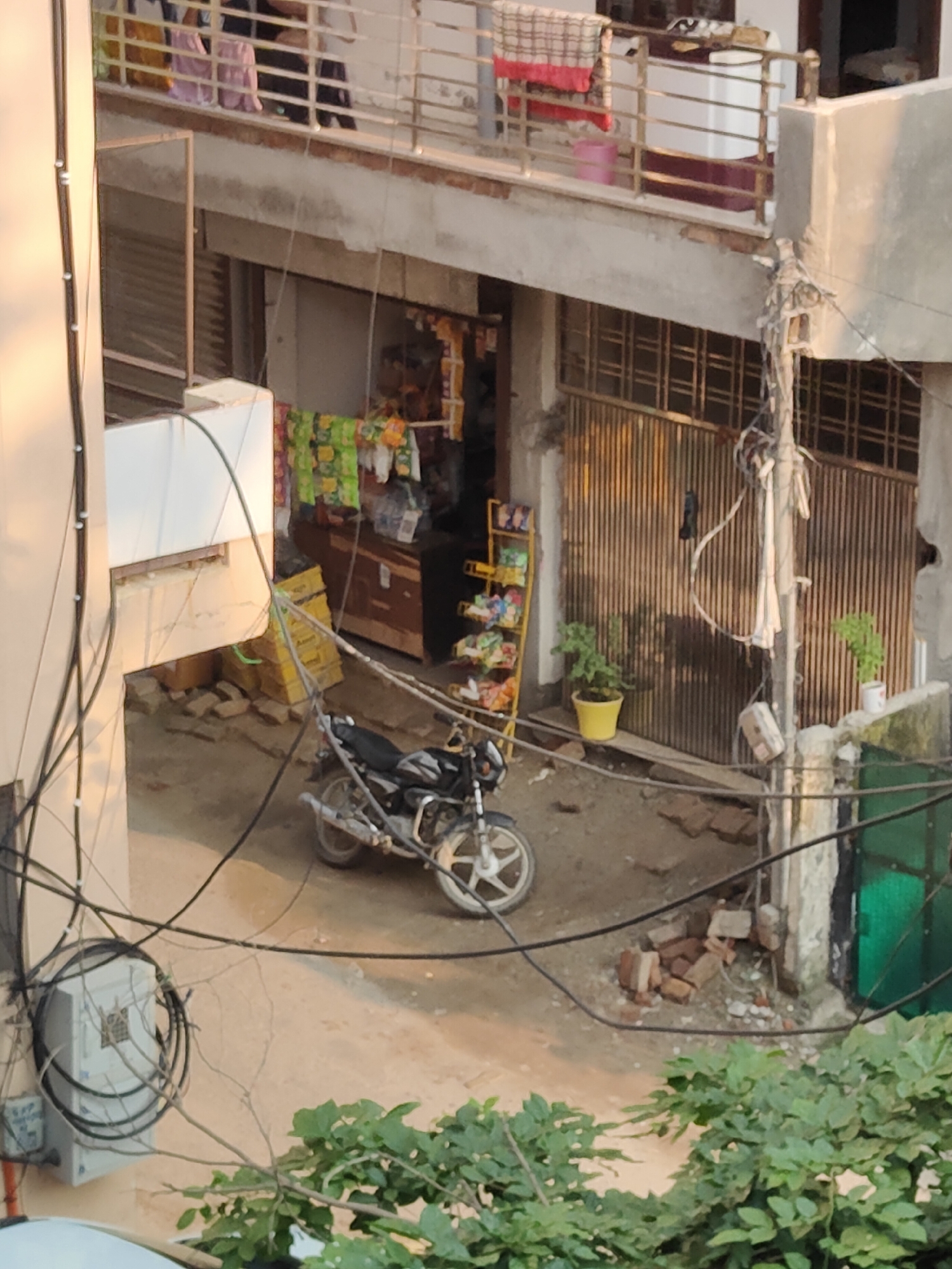 The image captures an outdoor scene in a narrow street or alleyway in Gurgaon, India, during daylight hours. In the foreground, a partially visible motorcycle is parked on a dirt ground. The motorcycle is black with white accents on the wheel. Behind the motorcycle, there's a shop or building entrance with a tall, ribbed metal gate. To the left of the gate, a yellow shelf unit displays numerous colorful packaged snacks. A small green plant in a yellow pot sits in front of the gate. To the right of the gate, another plant in a white pot is visible on a ledge. The building facade appears to be made of concrete and brick. Above the shop level, a second story is visible with a balcony railing. Laundry is hung on the balcony railing, including a pink bucket and fabric items. Several people are partially visible on the balcony, wearing casual clothing. The scene is cluttered with electrical wires and cables running across the frame. Some scattered bricks are present on the ground near the shop entrance. Lush green foliage is visible in the lower right and bottom edges of the image, suggesting the presence of trees or plants. The overall impression is one of a busy, somewhat disorganized urban environment.