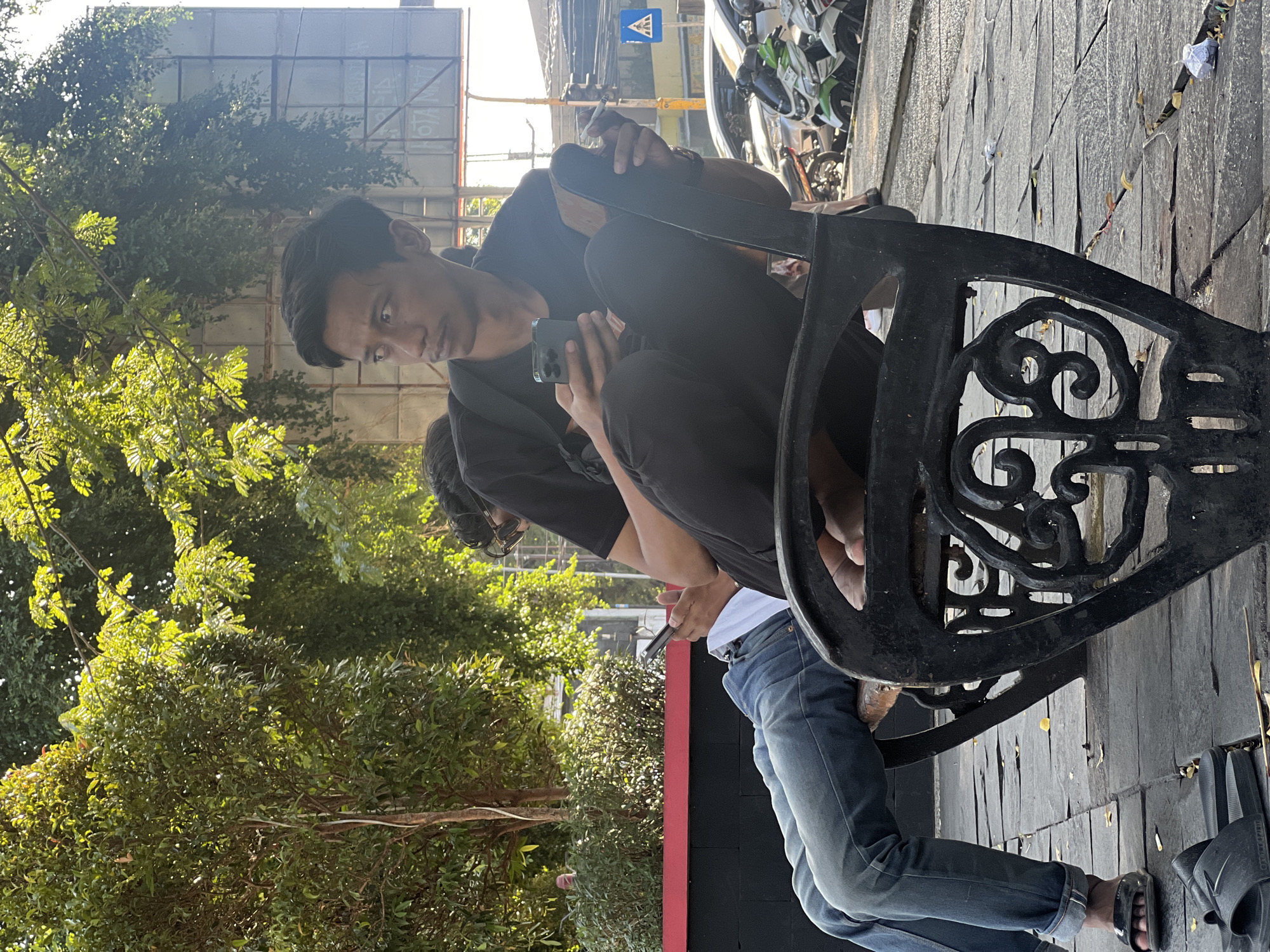 In Purwodadi, Indonesia, two individuals are seated on a black, ornate bench on a paved outdoor area. The person on the left, wearing blue jeans and black sandals, is leaning back with their phone in their lap and their legs extended. The person on the right, dressed in a black t-shirt and black pants, is seated upright, holding a smartphone in their left hand and a lit cigarette in their right hand. They are looking to their left. In the background, a large billboard stands partially obscured by lush green trees bathed in sunlight. The ground is paved with stone tiles and scattered with dried leaves. Parked motorcycles and cars are visible further in the background, suggesting a roadside or public space.