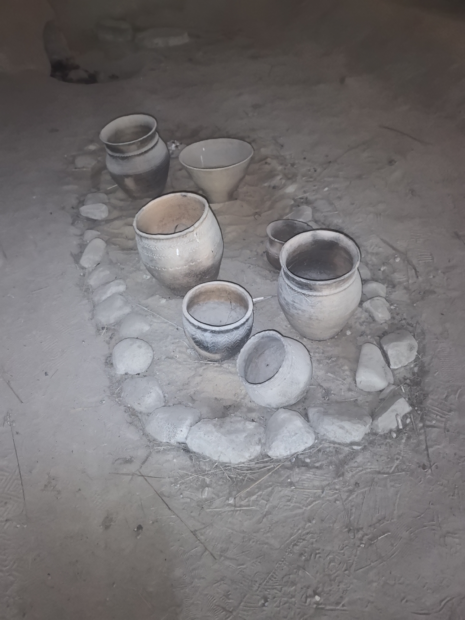 In Камянюцкі сельскі Савет, Belarus, a collection of ancient, earthenware pots and bowls are arranged in a circular pattern on a dirt floor. The pottery varies in size and design, with some exhibiting intricate etchings and decorative patterns. The pots appear to be aged, showing signs of wear and tear, with a rough, textured surface. The objects are placed on a floor made of compacted dirt, with several stones scattered around the pottery, possibly forming a border or support structure. The overall impression is that of an archaeological site or a display of historical artifacts, perhaps in a museum or an outdoor exhibit. The lighting is dim, casting subtle shadows and highlighting the textures of the pottery and the surrounding ground.
