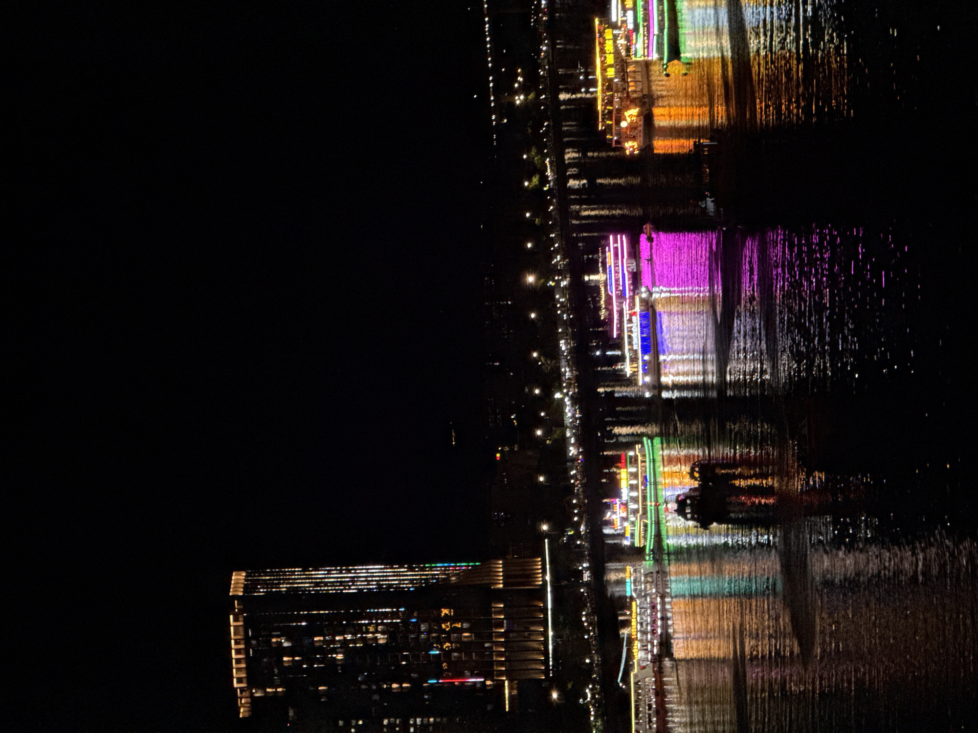 This is a nighttime scene in Da Nang, Vietnam, looking across the Han River at the illuminated Dragon Bridge. A tall, modern skyscraper on the left bank is brightly lit with vertical lines of lights and rows of horizontal yellow lights at the top. Several tour boats are docked or moving on the river, their decks and sides adorned with colorful LED lights in green, pink, and yellow. The lights from the buildings and boats are reflected on the dark, rippling surface of the water, creating streaks of color. In the foreground, a small dark boat with two occupants is visible, navigating the water. The background is dark, with scattered lights indicating more distant buildings and activity along the riverfront.