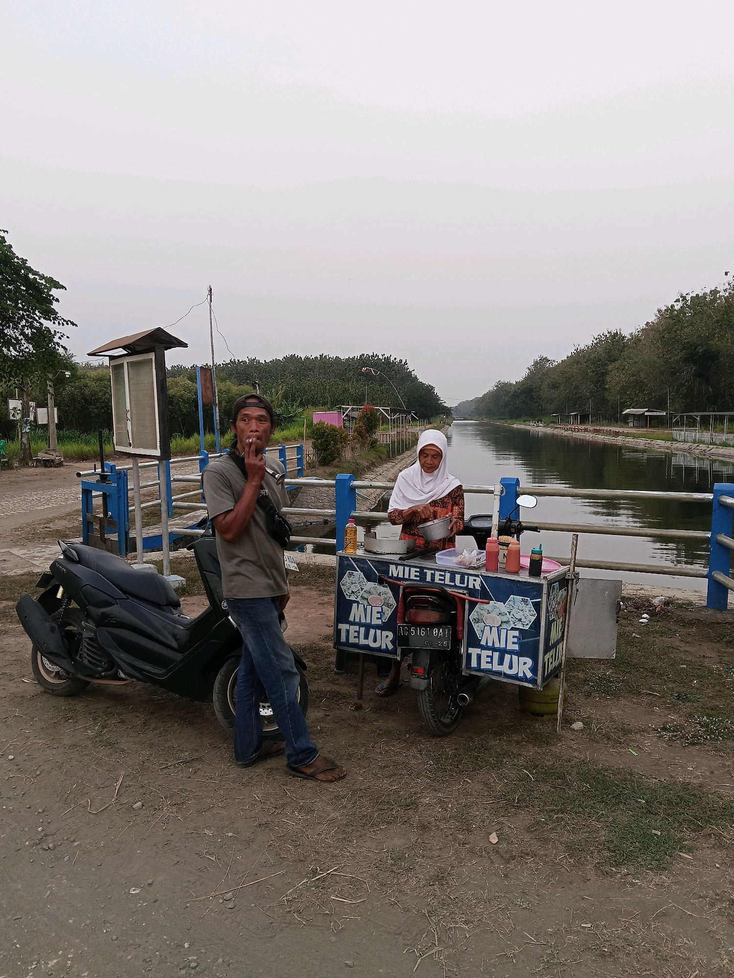 A man stands smoking next to a black motorcycle on a dirt path. He is wearing a gray t-shirt, blue jeans, a black baseball cap, and a dark fanny pack. To his right, a woman wearing a white hijab and a patterned dress is preparing food at a mobile food cart labeled "MIE TELUR". The cart is attached to a motorcycle. Behind them is a wide canal with trees lining the banks and a cloudy sky overhead. The scene appears to be located in Darungan, Indonesia, based on the signage and typical street food vendor setup.