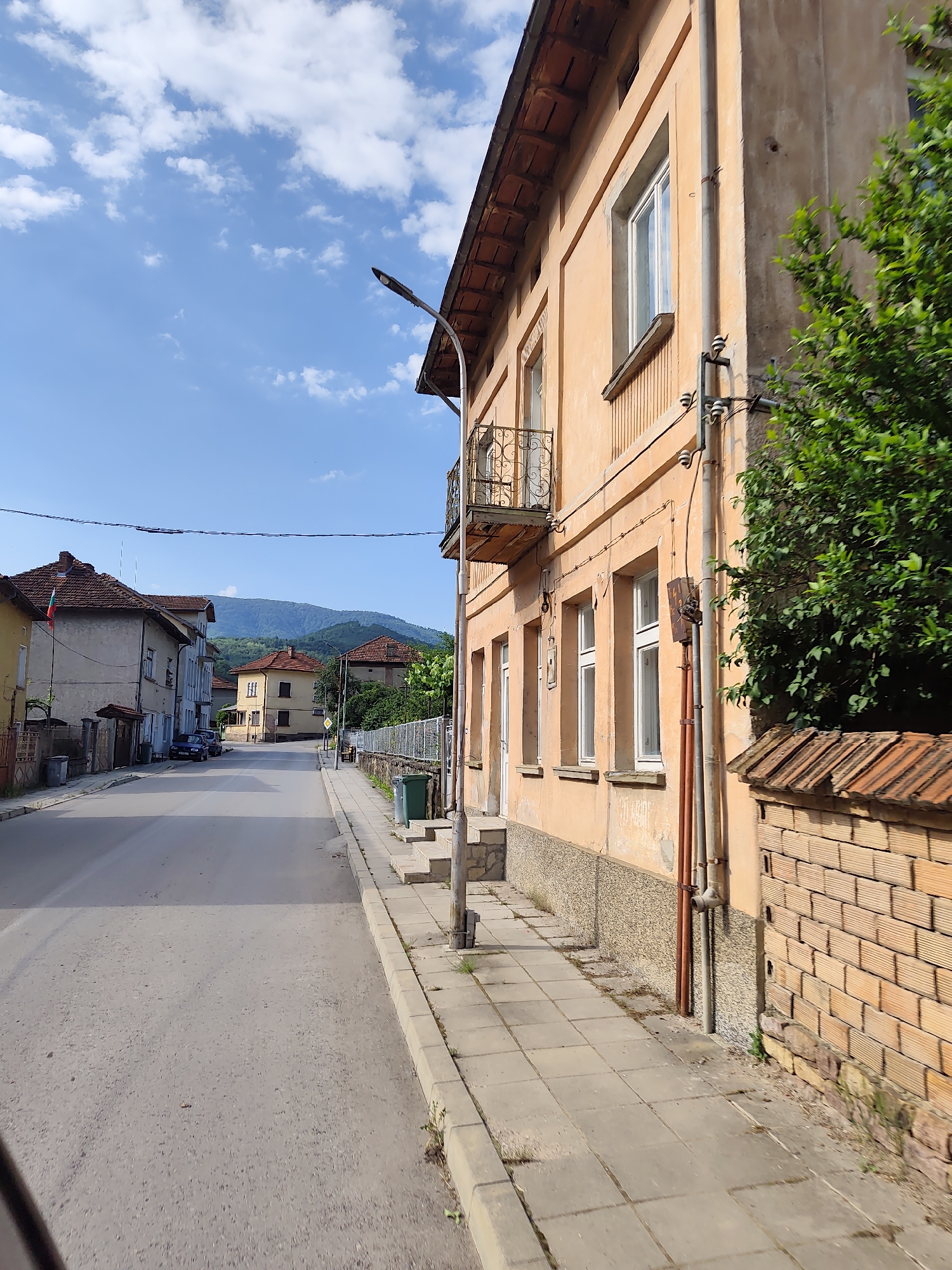 This is a street view in Lesidren, Bulgaria, on a sunny day with a bright blue sky and scattered white clouds. The perspective is from a vehicle looking down a paved street lined with buildings. On the right is a two-story building with an orange facade, featuring multiple windows, a small balcony with an ornate railing, and a tiled roof. A green tree grows next to this building, and a brick wall with a tiled top separates it from the sidewalk.
The street is empty of people but has parked cars visible in the distance. Buildings on the left side of the street are also two-story structures with tiled roofs. One building on the far left flies a Bulgarian flag. The street has a sidewalk on both sides, with some trash bins and a lamppost visible. In the background, rolling green hills form the horizon. The scene depicts a quiet, possibly rural, Bulgarian town.