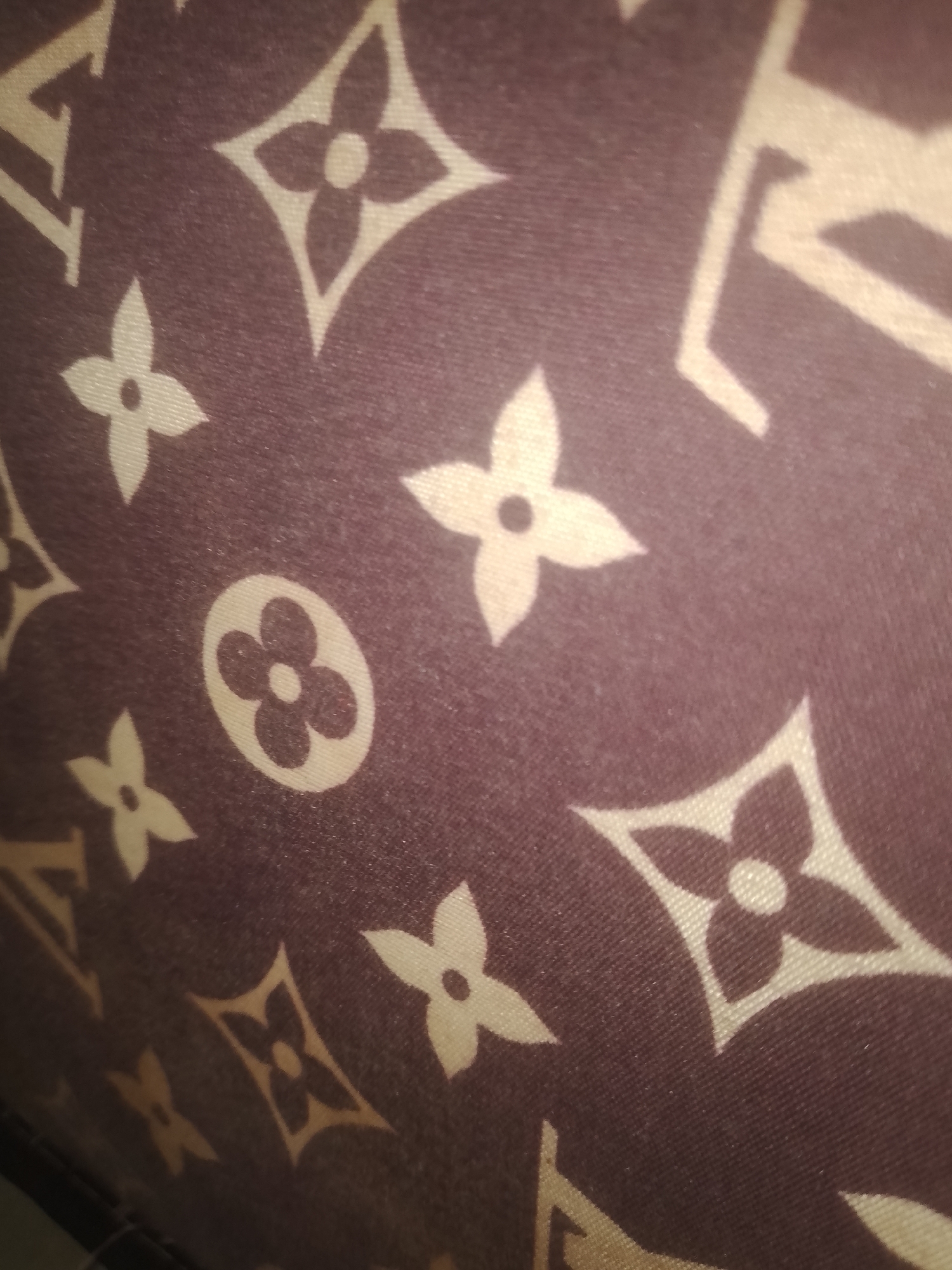 The image shows a close-up of a patterned fabric. The pattern consists of various floral and geometric shapes, in a light color against a dark brown background. Based on the pattern, it appears to be a Louis Vuitton monogram canvas. There is no information present in the image to suggest its location in Nkpor, Nigeria, or any other location. There are no people, actions, or interactions visible in the image.