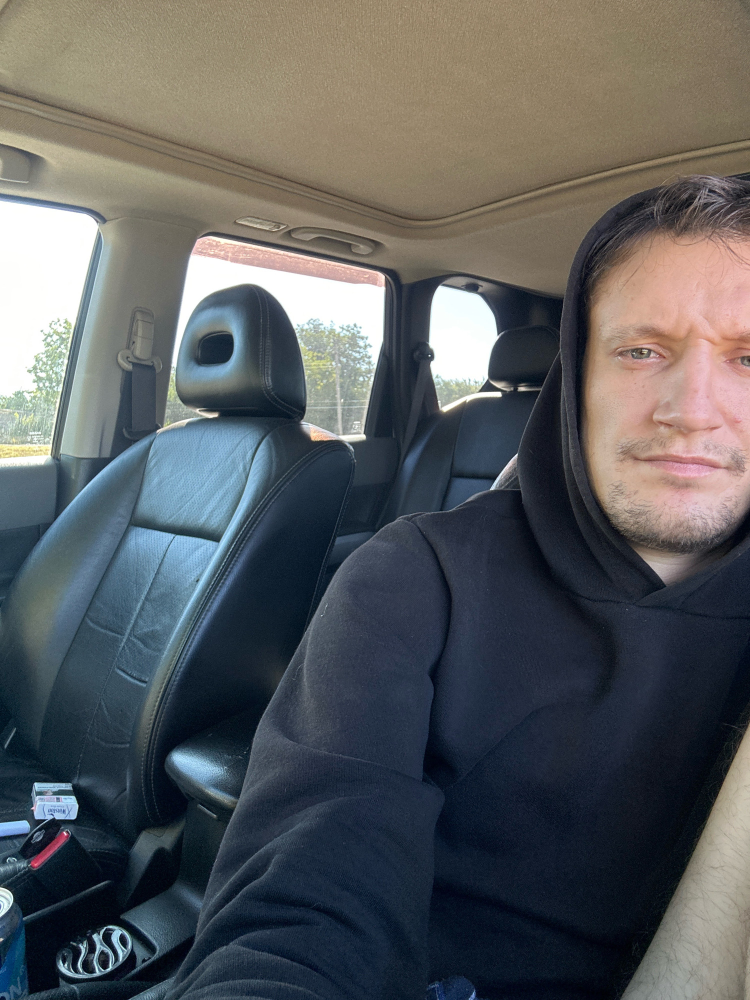 A man, appearing to be in his late 20s or early 30s, is seated in the driver's seat of a vehicle. He is wearing a black hooded sweatshirt. His gaze is directed towards the camera, and his facial expression is neutral. The interior of the vehicle is visible, featuring black leather seats and a sunroof. On the passenger seat, a pack of Winston cigarettes is placed next to a car key. A can, partially visible, rests in a cup holder. The background shows a landscape of trees and sky, suggesting an outdoor setting, possibly on the outskirts of Makiv, Ukraine. The lighting indicates it is daytime.