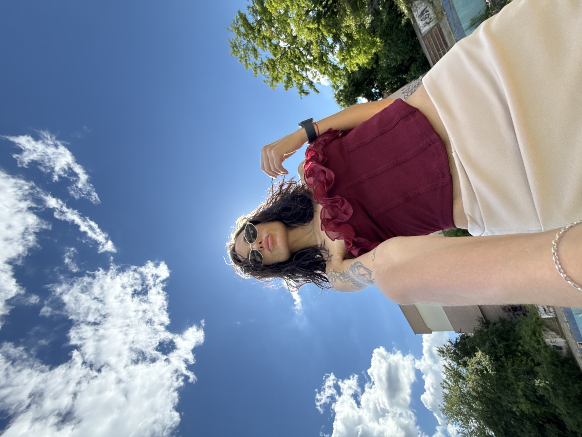 A low-angle shot captures a woman taking a selfie against a bright blue sky with scattered clouds. She is wearing a burgundy ruffled corset and a light-colored skirt. Her sunglasses are dark and angular, and her lips are pursed. Her arm is extended towards the camera, and a silver bracelet is visible on her wrist. Tattoos are present on her left arm. Green trees and a building with beige and brown siding are visible in the background, suggesting an urban environment. The sun is bright, casting a strong backlight. The location is identified as Pleven, Bulgaria.