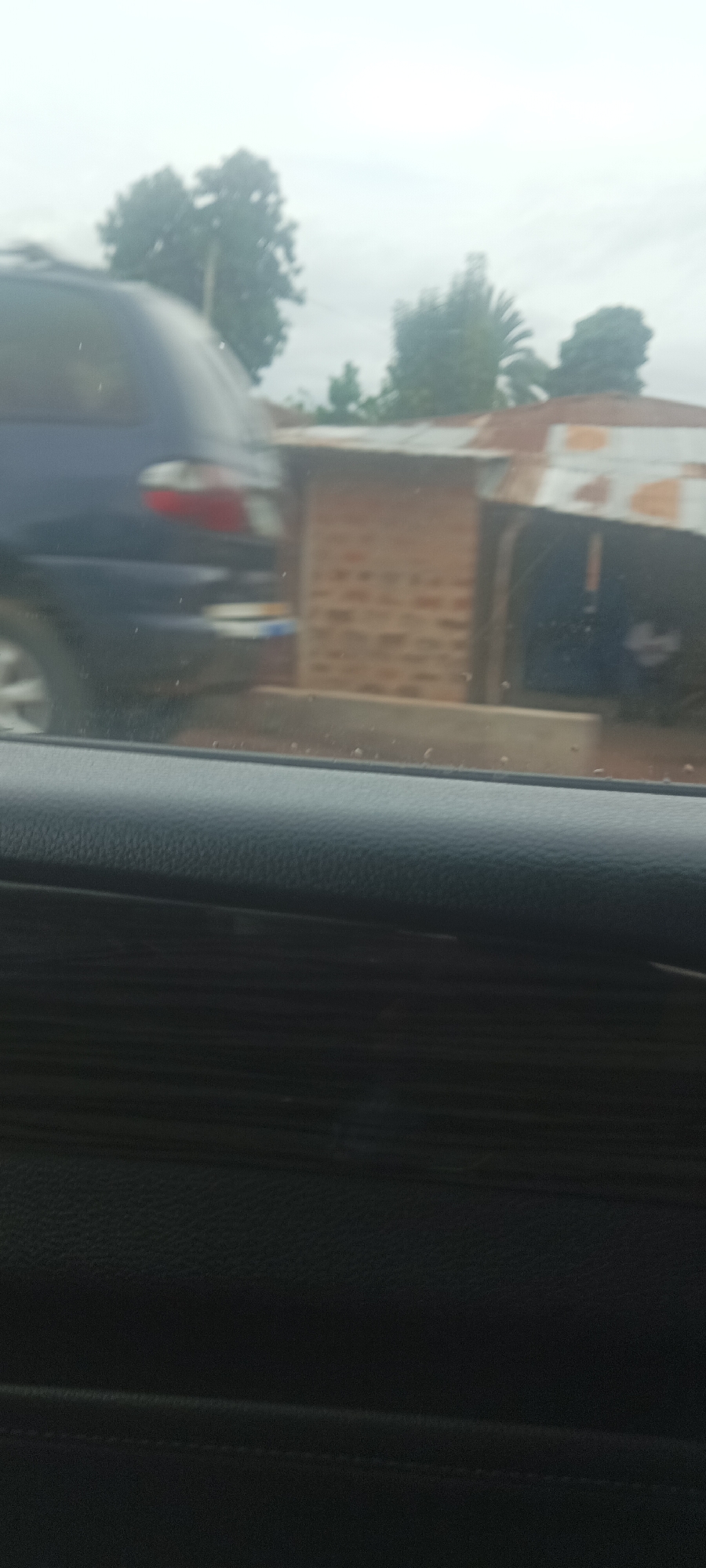 The image, likely taken from inside a moving vehicle, shows a blurred view of a blue vehicle and a small brick building with a corrugated metal roof. The building's construction appears basic. A concrete barrier is visible between the road and the building. Trees can be seen in the background against an overcast sky. The scene is suggestive of a roadside environment in a developing area. The location is Kadarko, Nigeria.