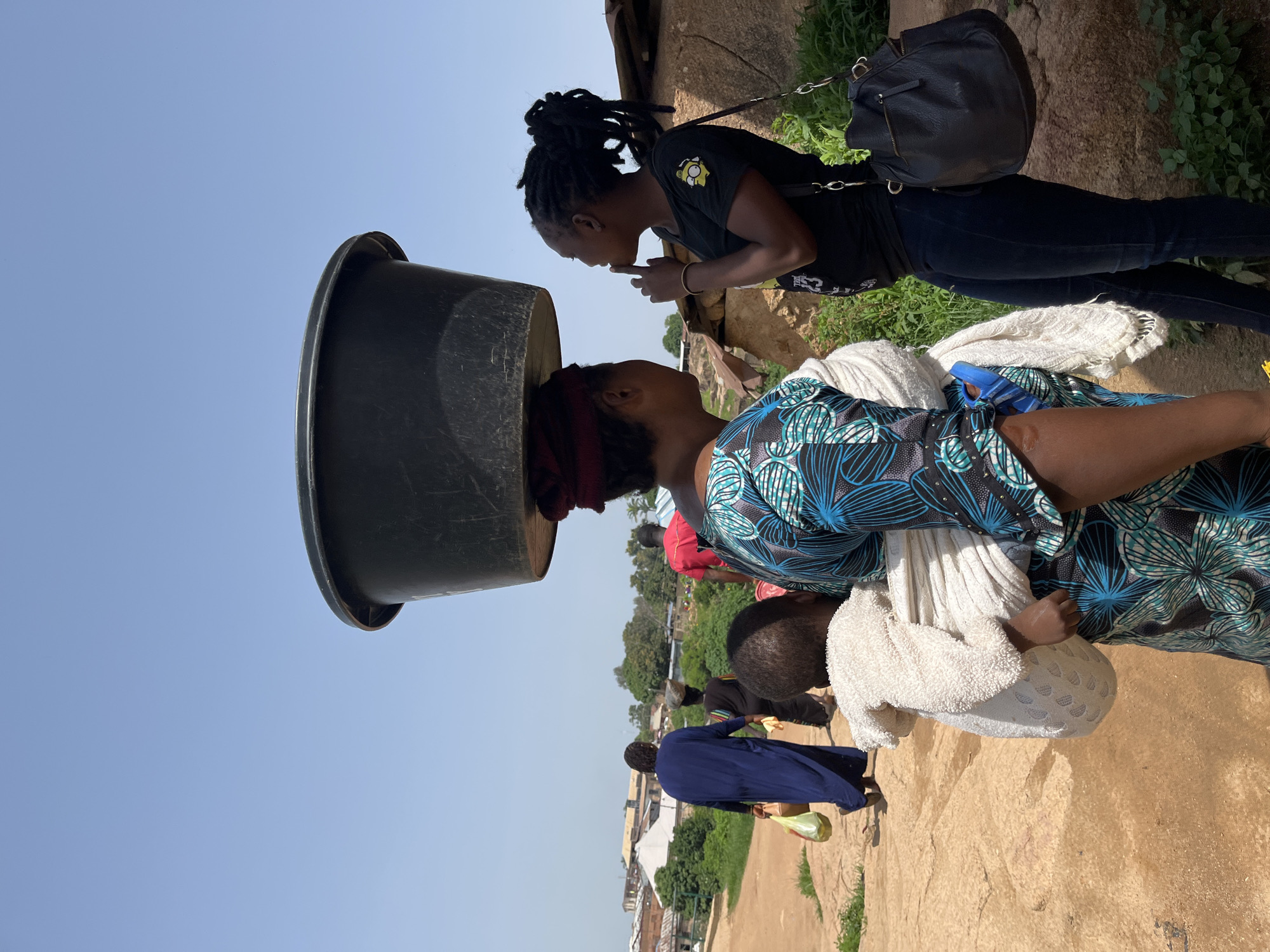 In the city of Jos, Nigeria, two women stand on a dirt path under a clear blue sky. The woman on the left carries a large black basin on her head, and her child is wrapped in a white cloth and slung across her back. She wears a blue and green patterned dress. The woman on the right, facing the first woman, wears a black t-shirt and dark pants and has her hair in braided dreadlocks. She holds a finger to her lips as if in thought or to silence. A black handbag with zippered pockets hangs from her shoulder. In the background, other people walk along the path, and buildings and vegetation are visible.