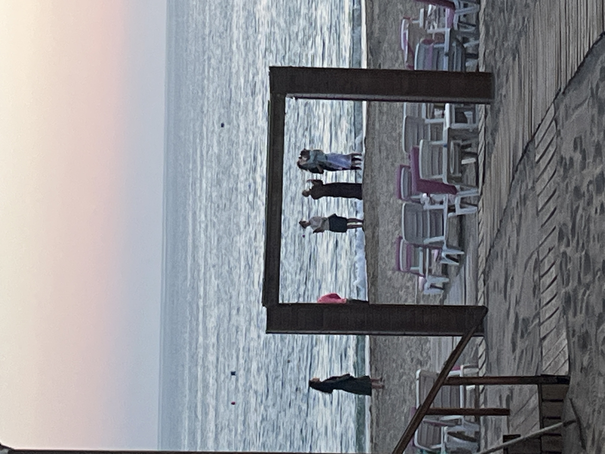 The image captures a serene coastal scene in Alanya, Turkey, likely during sunrise or sunset, indicated by the soft, diffused light and the pale, muted hues of the sky. The foreground and right side of the image are dominated by a sturdy wooden boardwalk or deck, suggesting a resort or promenade setting.
A large, dark wooden rectangular frame, possibly an architectural feature or a decorative archway, stands prominently between the boardwalk and the sea. Through this frame, several individuals are visible. One couple is positioned closely, appearing to embrace or stand in intimate conversation. Another person stands slightly apart within the frame, looking out towards the calm, rippling sea. Below the frame, closer to the bottom right of the image, another figure in dark attire stands alone on the boardwalk, also facing the water.
To the left of the boardwalk and around the wooden frame, numerous beach chairs with white frames and pink or grey seating are neatly arranged, indicating a leisure or relaxation area. The sea stretches into the distance under a hazy sky, with a few small, dark specks, possibly buoys or small boats, visible on its surface. The overall atmosphere is tranquil and contemplative, with the figures seemingly enjoying the peaceful coastal view. No visible text is present in the image.