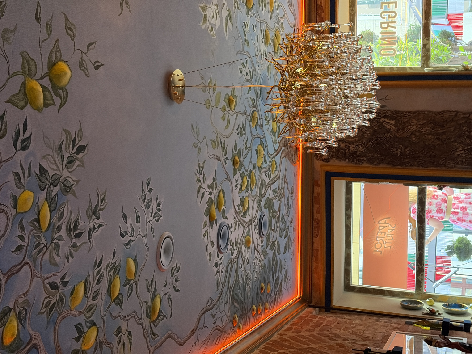 The image depicts the interior of a restaurant, possibly in Minsk, Belarus, with a distinctive ceiling mural of a lemon tree. The mural features branches and leaves painted in muted tones, with bright yellow lemons scattered throughout. A golden chandelier with numerous crystal teardrop-shaped accents hangs from the center of the ceiling, catching the light. Orange accent lighting runs along the edge of the ceiling where it meets the walls. The walls are partly brick, adding a rustic element. Through a window, a woman in a pink floral dress is visible, suggesting a public space. The window also has a sign with reversed Cyrillic text, likely indicating the name of the establishment. The overall atmosphere is warm and inviting, blending artistic decor with functional lighting.