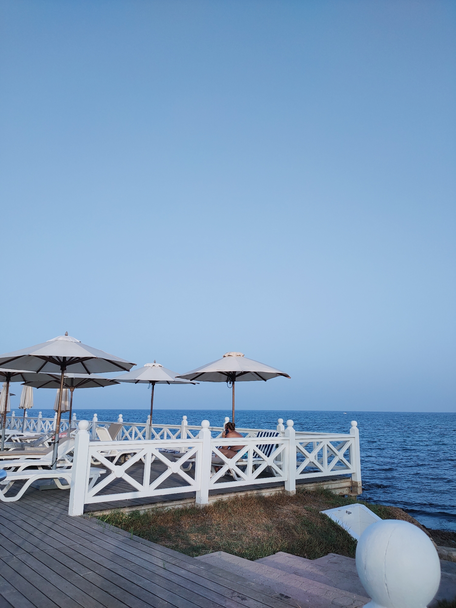 The image depicts a tranquil beach scene in Nabeul, Tunisia, under a clear blue sky. In the foreground, a wooden boardwalk leads to a white-latticed deck overlooking the calm, blue sea. Several white lounge chairs and umbrellas are arranged on the deck, suggesting a resort or public beach area. One person is visible on a lounge chair, partially obscured by a striped cushion, facing away from the camera. The environment is serene, with the gentle lapping of waves against the shore and the vast expanse of the sky contributing to a peaceful atmosphere.