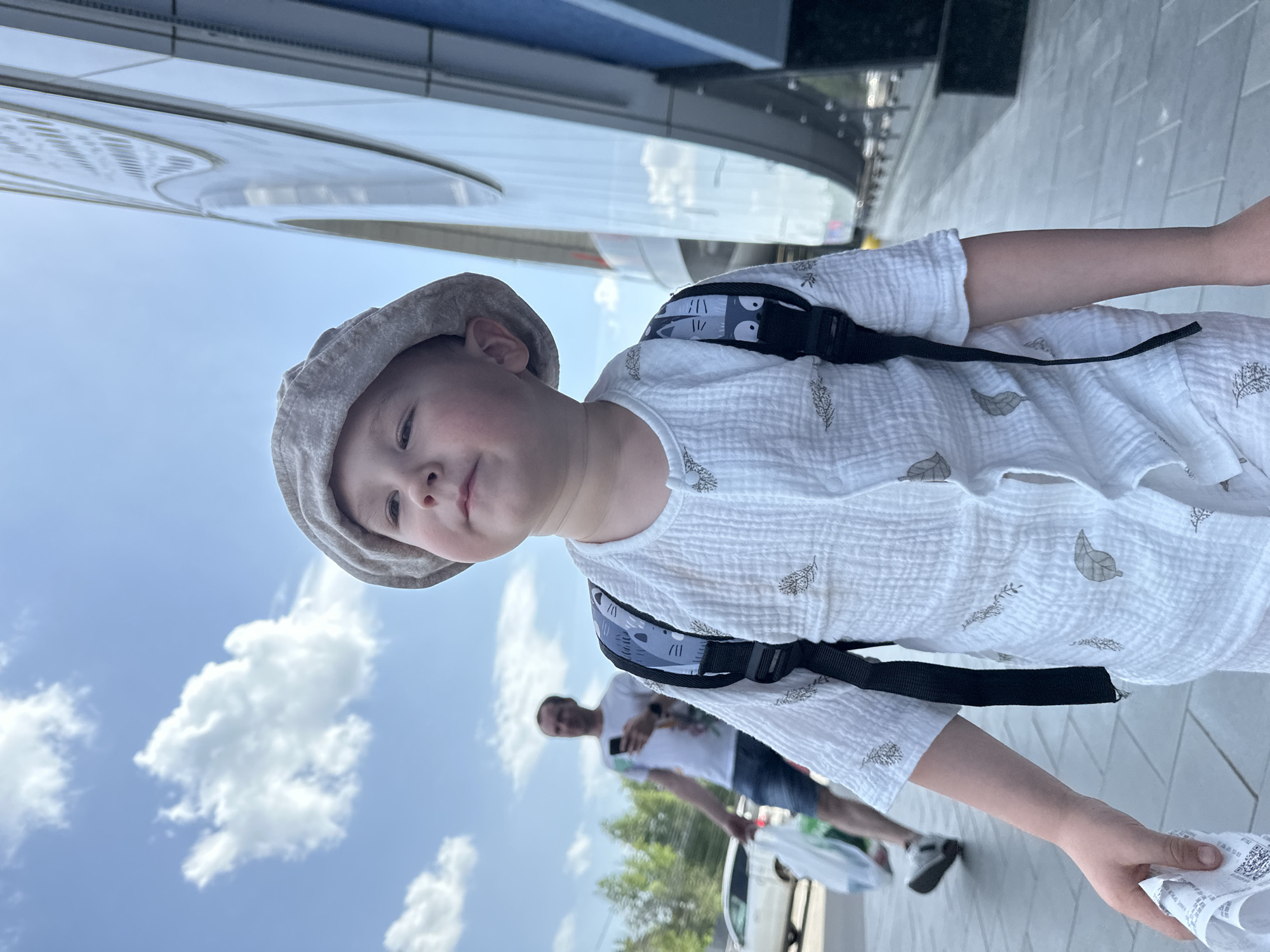 A child wearing a beige beret, a white short-sleeved shirt with a foliage pattern, and a dark-colored backpack stands near a reflective building with a curved exterior, on a light-colored tiled surface. The sky behind the building is blue with fluffy white clouds. An adult with a white T-shirt and blue shorts is in the background, slightly blurred. A white car is visible at the bottom of the frame. The scene appears to be in an urban environment. The photograph was taken in Nizhny Novgorod, Russia.