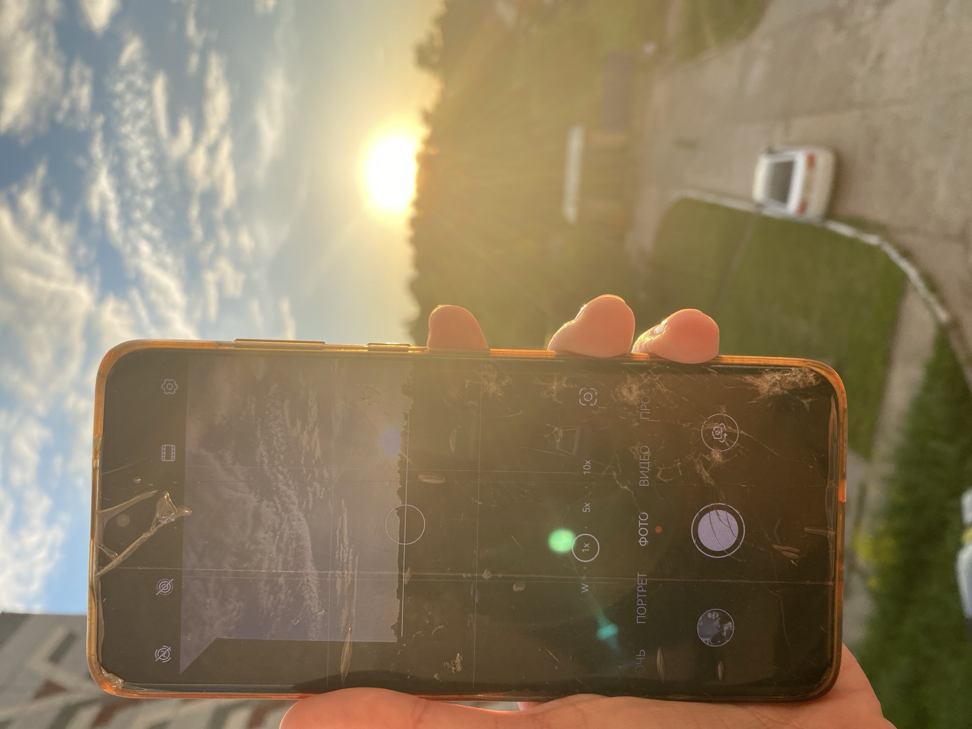 A human hand, with visible fingers, grips the left side of a smartphone, prominently displayed in the foreground. The phone's screen is severely cracked and scratched, showing numerous linear fractures and surface abrasions, particularly concentrated at the top near what appears to be the front camera notch. The device is enclosed in an orange-colored protective case. The phone's display shows a camera application interface, with "ФОТО" (Photo) mode actively selected, indicated by a red dot. Zoom options "W", "1x", "5x", and "10x" are visible, with "1x" highlighted. Other language-specific mode selections include "ПОРТРЕТ" (Portrait), "ВИДЕО" (Video), and "ПРОФ" (Pro). The camera preview on the phone's screen displays a sky with scattered clouds and a dark, distant horizon, captured under a significantly different light level than the surrounding environment. Green and blue lens flares are visible on the phone's screen, indicating strong light exposure.
The background, rendered out of focus, depicts an outdoor scene at sunset in the city of Chaikovsky, Russia. A brilliant, golden sun, positioned just above a distant treeline, dominates the upper right, casting a warm, luminous glow across the entire scene. The horizon is defined by a dense expanse of dark trees. Below the treeline, a paved path or road curves from the lower right towards the center. A white sedan is parked on the right side of this paved area. To the left of the path, green grass is visible, bordered by a white curb or railing. In the mid-distance, a blurred, light-colored structure, possibly a small pavilion or shelter, is discernible. On the far left, a section of a multi-story residential building with angular architectural features is partially visible against the sky, which features scattered, wispy clouds.