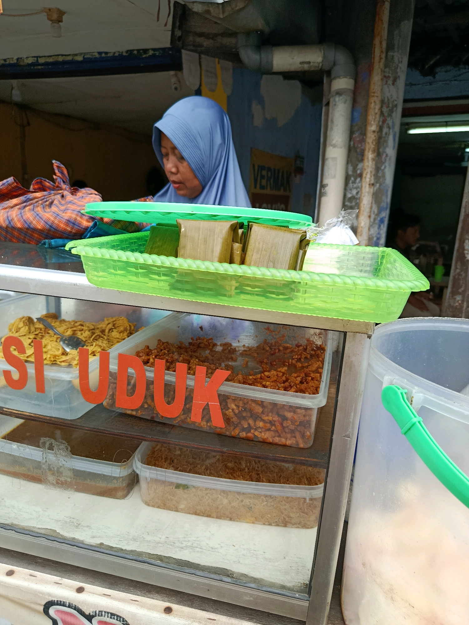 In Bekasi, Indonesia, a street vendor in a blue hijab displays food for sale. In the foreground, a glass display case contains several dishes. The most prominent is a clear plastic container with orange lettering spelling "SI UDUK", which holds a portion of fried tempeh seasoned with spices. To the left of this, a metal spoon rests in a dish containing a yellow, shredded food item, possibly fried noodles or shredded chicken. Below these, two more clear plastic containers hold what appears to be a dark brown sauce and a pile of fried shallots. Above the display case, a green plastic basket holds several portions of food wrapped in banana leaves, some of which are square-shaped. The vendor, with her head and shoulders visible, is looking down at the food. In the background, pipes and peeling paint are visible on a wall, and a yellow sign with the word "VERMAK" is partially obscured. The overall scene suggests a bustling street food stall.