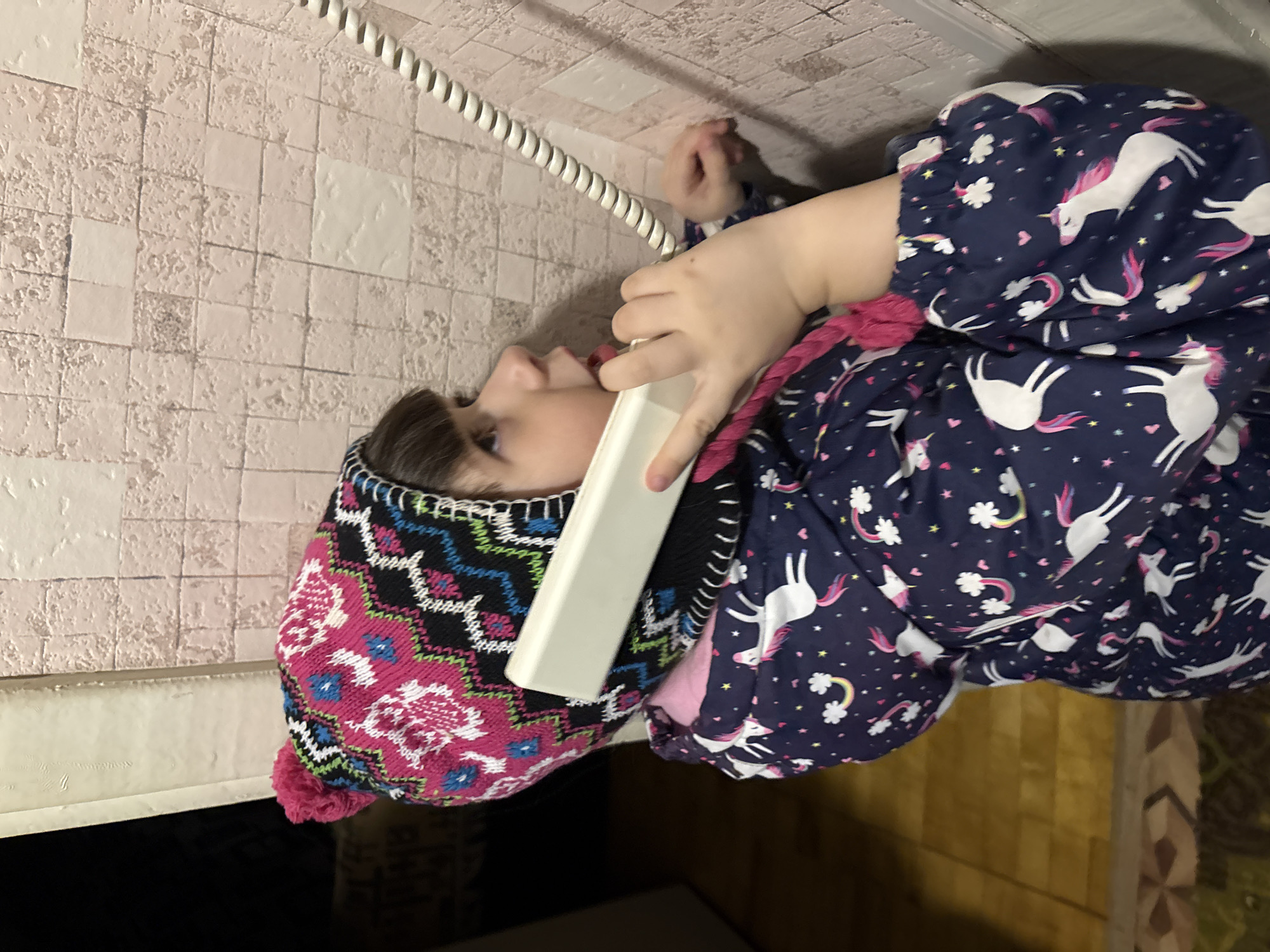 A young child is depicted in an indoor setting in the city of Chaikovsky, Russia. The child, with dark hair and bangs, wears a multi-colored knitted winter hat with a fuchsia pom-pom and a geometric pattern in fuchsia, black, white, blue, and green. She is dressed in a navy blue puffer jacket patterned with white unicorns, pink hearts, white clouds, and rainbows. The child holds a cream-colored landline telephone receiver to her left ear with her left hand, her mouth slightly open, and her gaze directed upwards and to the right. A coiled white telephone cord extends upwards from the receiver, running against a light-colored wall with square, textured tiles. A dark doorway is partially visible in the left background, and a section of tiled floor with brown and patterned tiles is visible at the bottom left.