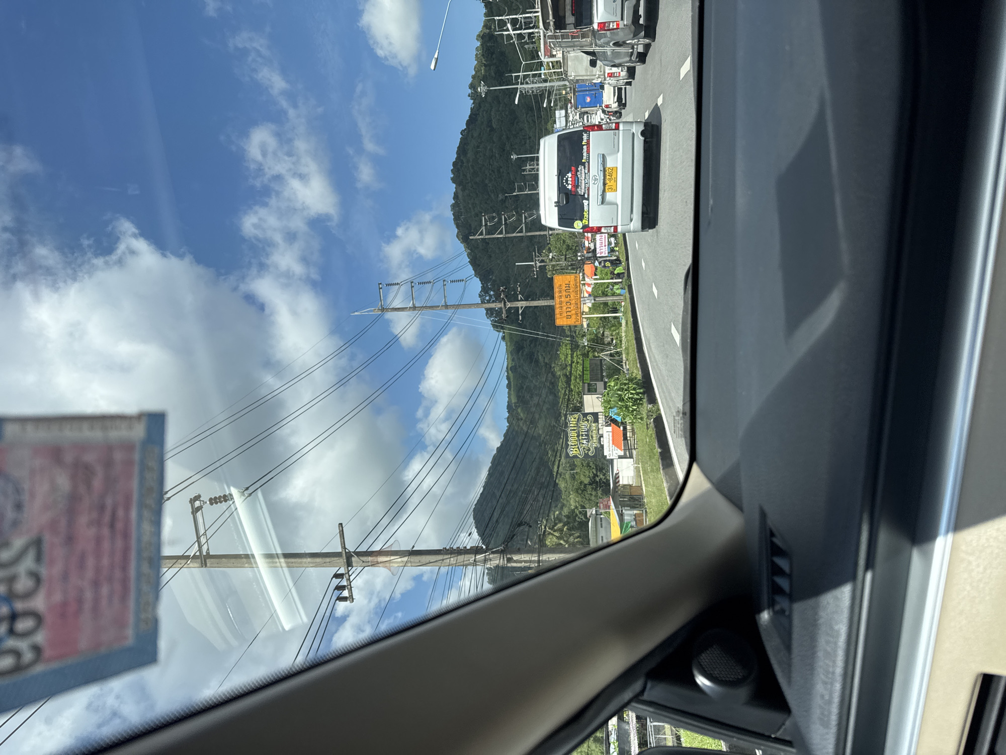 The image shows a view from inside a vehicle traveling on a road in Kathu, Thailand. The road is congested with traffic, including a silver van with a license plate "31-8462" in front of the vehicle. On the right side of the road, there is a billboard for "Bloodline Tattoo." Further ahead, there are more vehicles and buildings. The sky is blue with some white clouds, and there are mountains in the background. Power lines are visible overhead.
