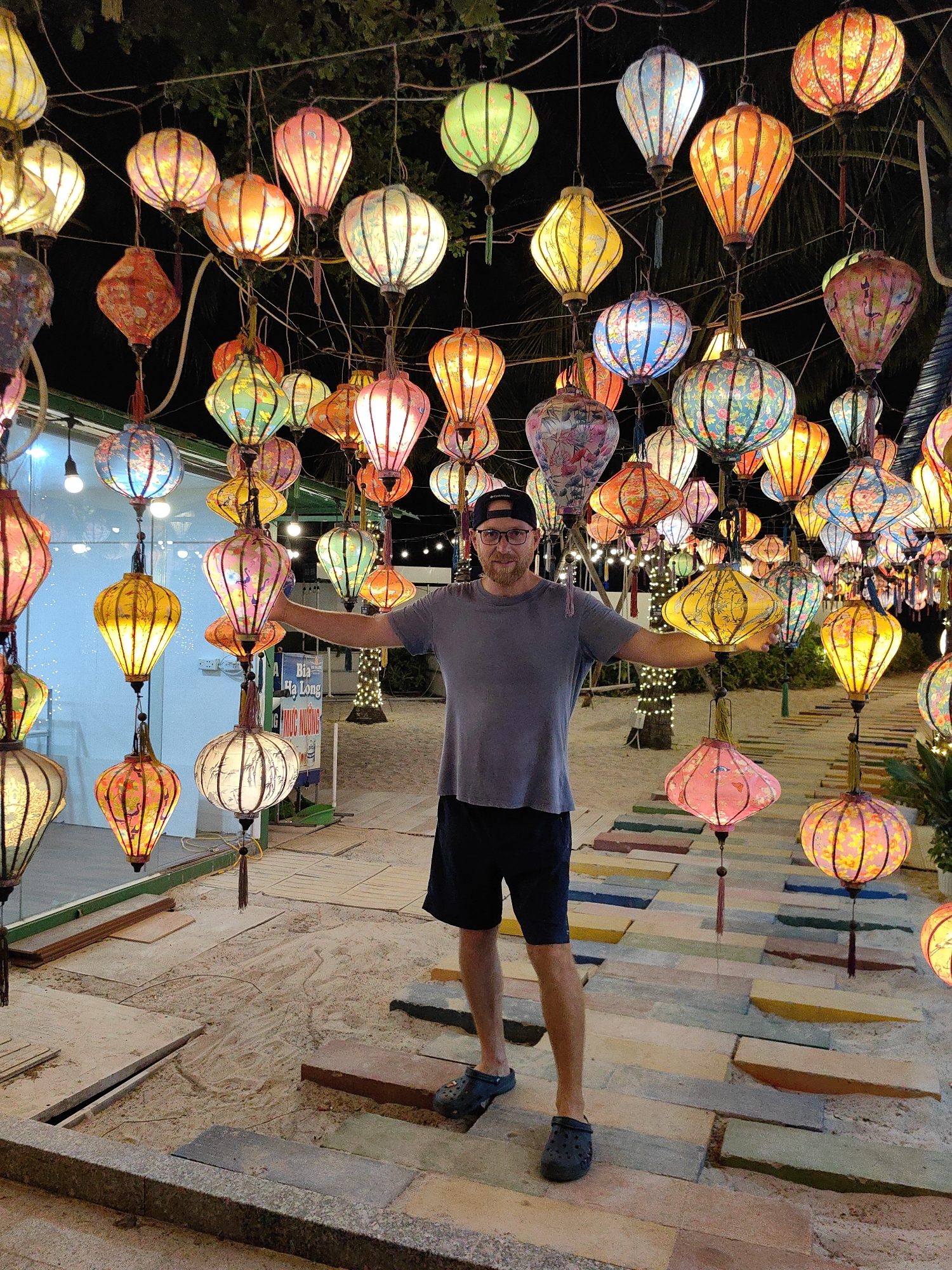A man stands with outstretched arms amidst a display of vibrant lanterns in Bai Chay Ward, Vietnam. The man, wearing a grey t-shirt, black shorts, a black baseball cap, and dark-colored clogs, is the central figure. He is positioned on a pathway made of multicolored rectangular paving stones, which are partially covered in sand. Above him, numerous colorful lanterns hang from wires, creating a canopy of light. In the background, signs, trees, and other vegetation are visible. The scene is illuminated by the light emanating from the lanterns and other ambient light sources.