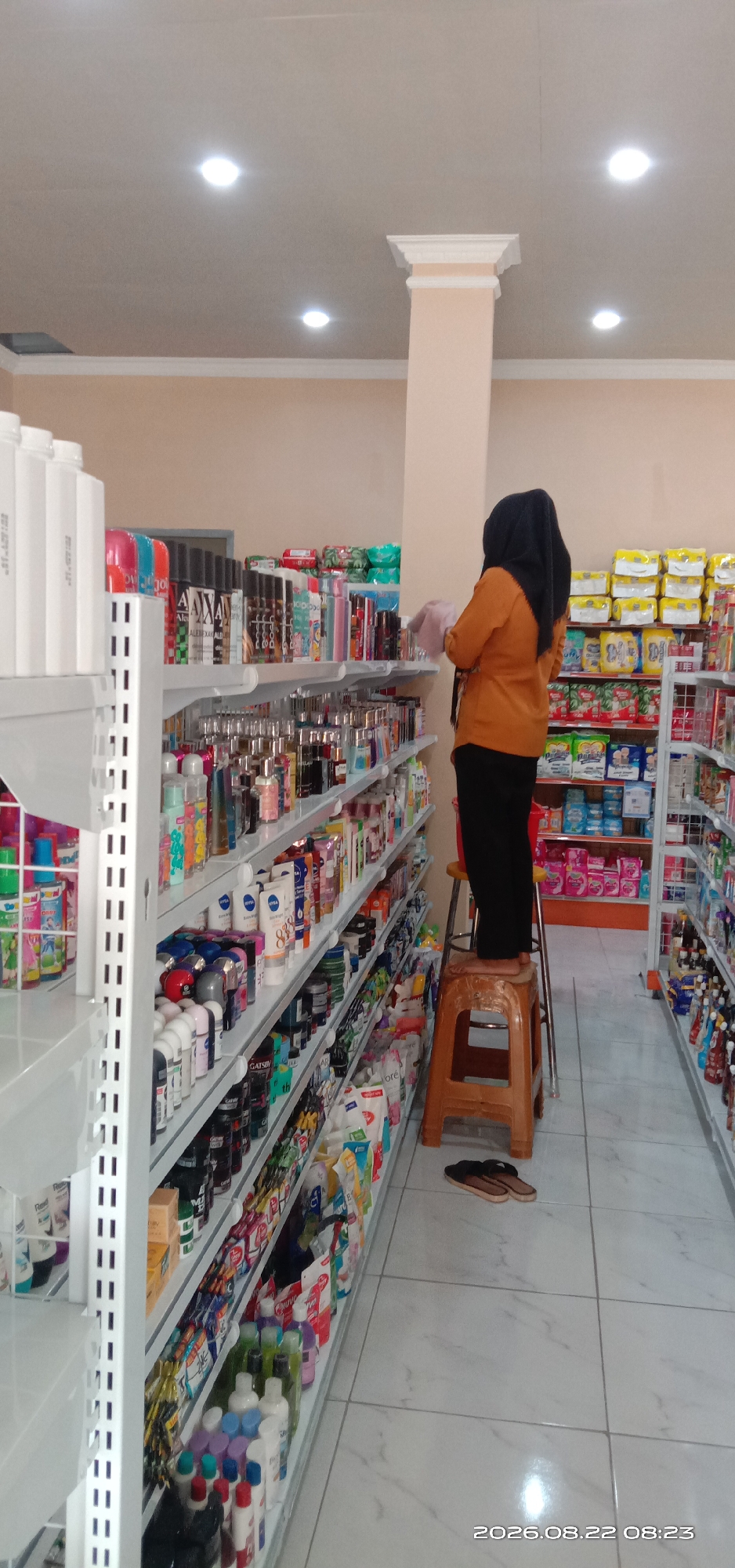The image depicts the interior of a retail store, likely a minimarket or a pharmacy, located in Pakong, Indonesia. A woman wearing an orange shirt and a black hijab stands on a small, brown plastic stool, reaching for a product on a high shelf. She is holding a light pink cloth in her right hand, suggesting she might be cleaning or organizing the merchandise.
The store features multiple rows of shelves stocked with a variety of products. The shelves on the left are filled with toiletries, cosmetics, and possibly personal care items, presented in various bottles and packages. The shelves on the right display packaged goods, including what appear to be detergents or cleaning supplies, and possibly food items. The overall impression is one of a well-organized retail environment. The floor is tiled with a light-colored pattern. A prominent light-colored pillar divides the space. A pair of brown and black flip-flops are placed on the floor near the stool. The date stamp on the image indicates "2026.08.22 08:23".