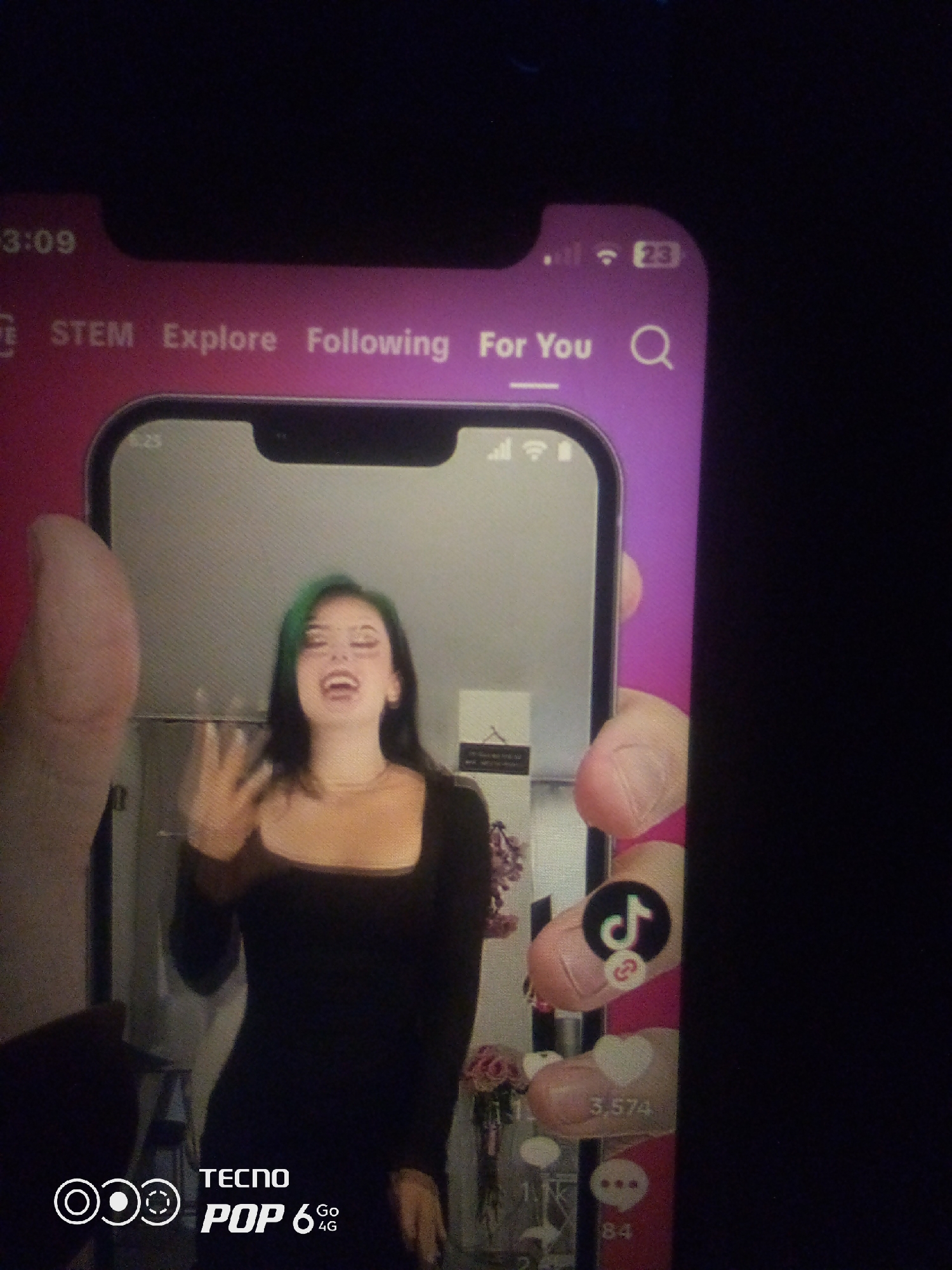 The image is a photograph of a smartphone screen displaying a social media application, likely TikTok, held by a person whose hand is visible on the left side. The phone screen shows a woman with green-streaked black hair, wearing a black dress, with her mouth open and her left hand raised in a gesture. The interface of the app displays icons for likes, comments, and shares, along with numerical counts. The phone's status bar at the top shows the time as 13:09, battery indicator, and network signal strength. The background of the phone's display appears to be an indoor setting, possibly a room with some decorative items like flowers visible. The phone itself has a bezel and notch characteristic of modern smartphones. The phone is a TECNO POP 6 4G. The surrounding environment is dark, suggesting the photograph was taken in low light conditions, and no specific details about Howayi, Nigeria, are discernible from the image itself.