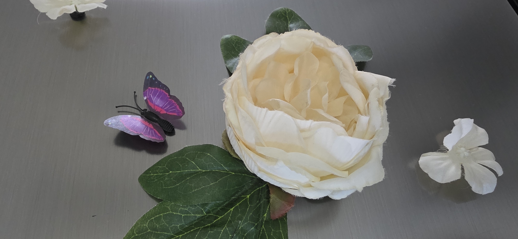 A vertical close-up shot features a collection of artificial floral decorations and a decorative butterfly against a neutral, grey background. In the upper right quadrant, a cluster of delicate white petals, likely from another artificial flower, is partially visible, casting a soft shadow. Below this, to the left, a vibrant, iridescent purple and pink butterfly is positioned, its wings slightly ajar, with black antennae. Dominating the lower half of the image is a large, creamy yellow artificial rose, its petals soft and layered, with two green leaves extending from its base. To the lower left of the rose, a single, small, white artificial flower with five petals lies detached. The background appears to be a smooth, matte surface, possibly a tabletop or wall. There are no people or animals present, and the setting is devoid of any specific city or geographical markers, thus it cannot be placed within Sumgayit, Azerbaijan. The lighting is soft and even, highlighting the textures and colors of the decorative items.