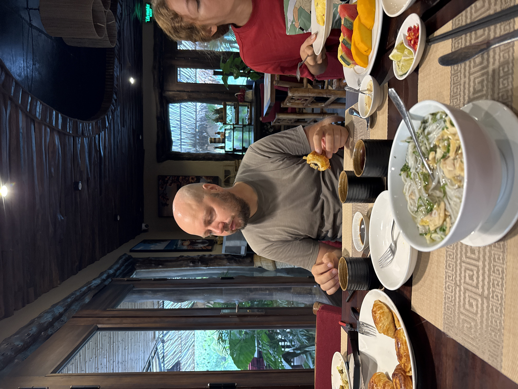 A man and a woman are seated at a wooden table in a restaurant in Nam Hoa Lư Ward, Vietnam, enjoying a meal. The man, with a bald head and a beard, wears a grey t-shirt and is holding a piece of food, appearing to be about to eat it. The woman, with curly brown hair, is wearing a red t-shirt and is in the process of eating from her plate.
The table is set with several dishes, including a bowl of pho, a plate of pastries, and a fruit platter with watermelon and mango slices. There are also small bowls of sauce and condiments, as well as cups and utensils. The restaurant's interior features a rustic design with wooden accents and exposed beams on the ceiling. Large windows look out onto a lush green outdoor area, suggesting a natural or garden setting. The lighting is dim, creating a warm and inviting atmosphere.
