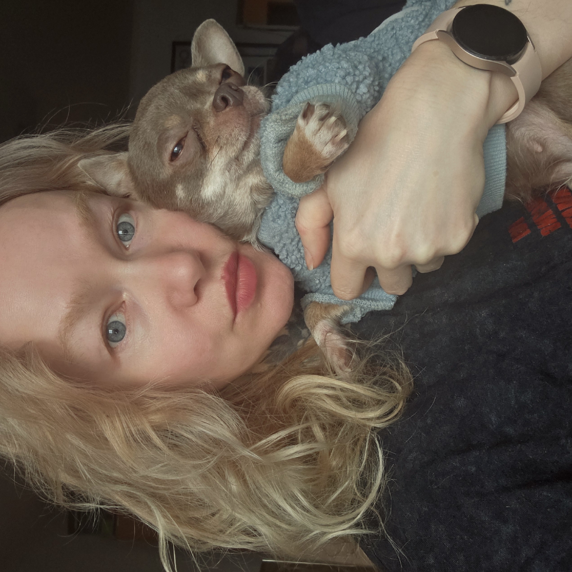 A fair-skinned woman with blonde hair and blue eyes is holding a small, light brown Chihuahua dog. The dog is wearing a light blue sweater. The woman is wearing a dark gray shirt and a pink smartwatch. The background is blurry and indistinct. The location is not identifiable as Saint Petersburg, Russia from the available visual information.