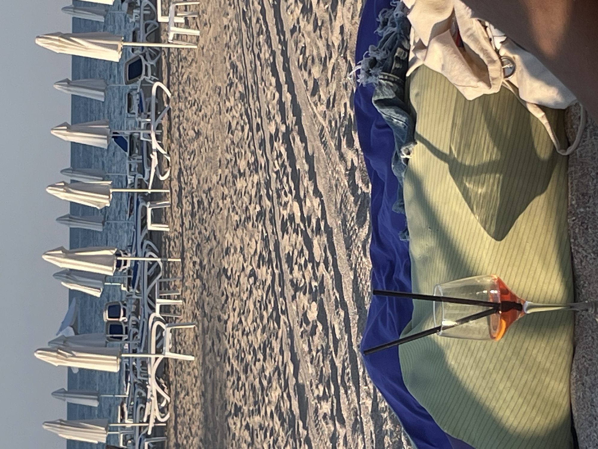 The image depicts a beach scene with rows of white lounge chairs and umbrellas set up on the sand. The chairs are neatly arranged, facing the ocean, with some appearing to be occupied by people from the clothing draped over them. The sand shows the texture of footprints or tracks, suggesting recent activity.
In the foreground, a partially visible person's arm and hand are resting on a colorful beach towel, which is placed on the sand. The towel is a vibrant blue and green, with some light-colored fabric on top, possibly clothing. A stemmed glass filled with an orange liquid and two black straws is prominently placed on the green towel. The glass is partially shadowed, and the orange drink suggests a refreshing beverage.
The lighting indicates it is either early morning or late afternoon, with soft shadows cast across the scene. The sky is a pale, hazy color, suggesting clear or slightly overcast weather. The overall mood of the image is relaxed and leisurely, typical of a beach vacation. The presence of the lounge chairs and umbrellas implies a resort or a designated beach area. There is no visible text in the image.