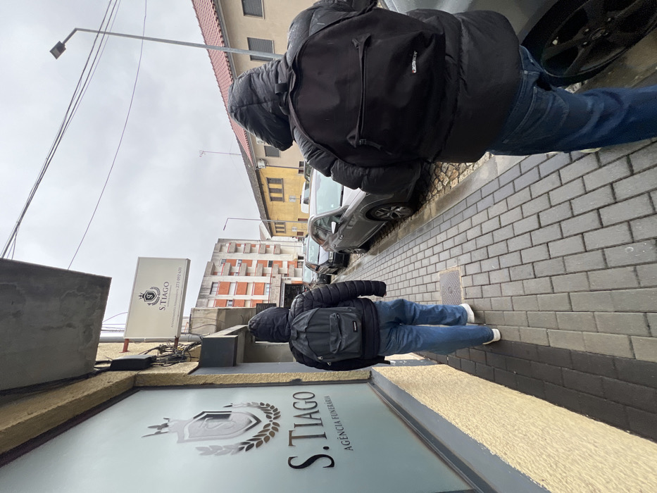 The image captures an outdoor urban scene on a cloudy day, identified as R. Trindade 134, Bragança, Portugal. The perspective is from behind two individuals walking away from the camera on a grey paver sidewalk.
On the left, a building with a textured yellow facade houses "S. TIAGO AGÊNCIA FUNERÁRIA," indicated by prominent signage on a large window and an illuminated rectangular sign above it. The sign displays a crest, "S. TIAGO," "ATENDIMENTO PERMANENTE," and the phone number "273 092 631."
The two individuals are both dressed in dark hooded puffer jackets, blue jeans, and black backpacks. One backpack exhibits an "Eastpak" logo. The individual on the left is wearing white-soled shoes.
To the right of the sidewalk is a street with multiple parked vehicles, including a grey SUV-like car in the midground. Across the street, a low-rise yellow building is visible, followed by a taller, multi-story residential building with orange balcony details in the background. Overhead wires and streetlights are present against an overcast sky. The sidewalk appears damp. No direct interactions between the individuals or with other subjects are observed.