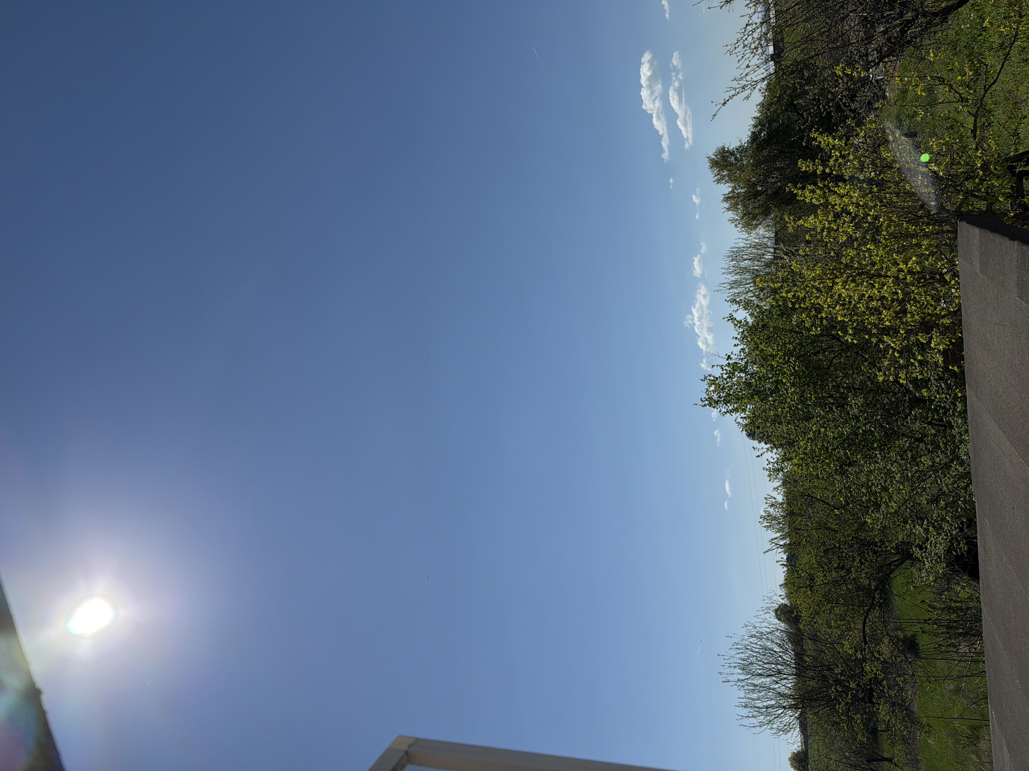 The image depicts a daytime scene with a clear blue sky, a bright sun, and trees. Thin clouds are visible near the horizon. The location is potentially Rosen, Bulgaria as requested, but that is not verifiable from the image alone. The scene includes elements from nature with trees lining the right side and the sun. A portion of a building structure is visible in the bottom left and bottom center of the frame.