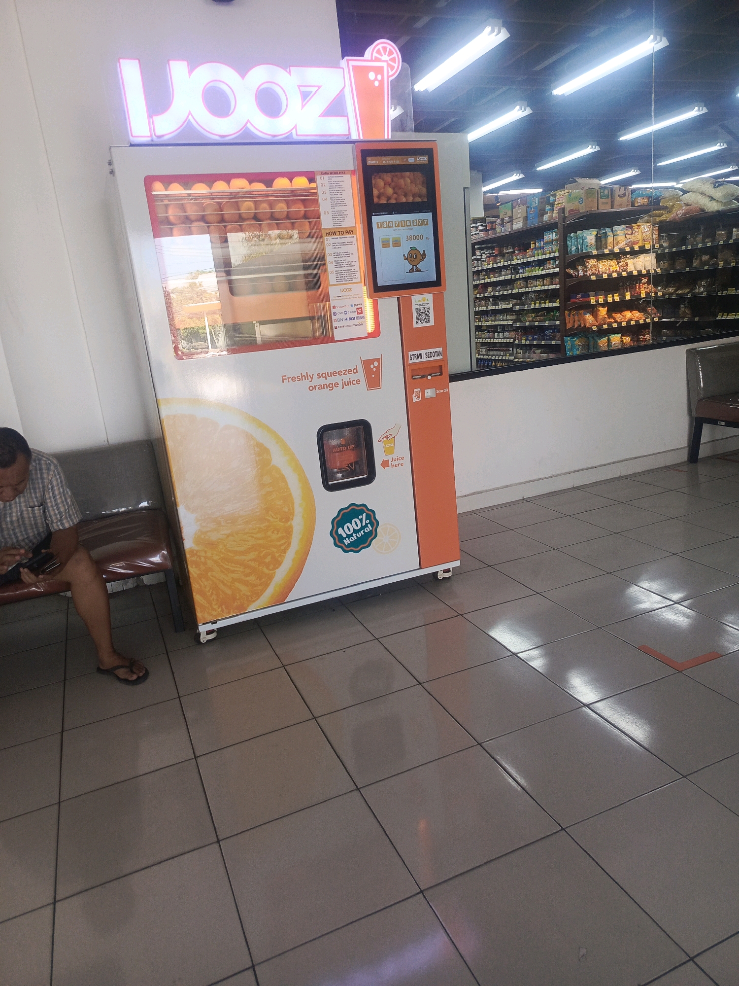 The image depicts a "JOOZ" branded fresh orange juice vending machine located in a convenience store in North Kuta, Indonesia. The machine, predominantly orange and white, displays a large graphic of a halved orange and the text "Freshly squeezed orange juice" and "100% Natural". A screen on the machine shows a price of 38000 Rp and a cartoon orange character. Instructions for use and payment options, including various e-payment providers like ShopeePay and Gopay, are visible on the side panel. A dispenser for straws is also present. The machine is positioned on a tiled floor, and to the left, a man in a plaid shirt and flip-flops is seated on a bench, looking at his phone. The background reveals shelves stocked with various products, typical of a convenience store or minimarket, with fluorescent lighting illuminating the scene. An orange directional arrow sticker is visible on the floor in the background, suggesting a queuing system.