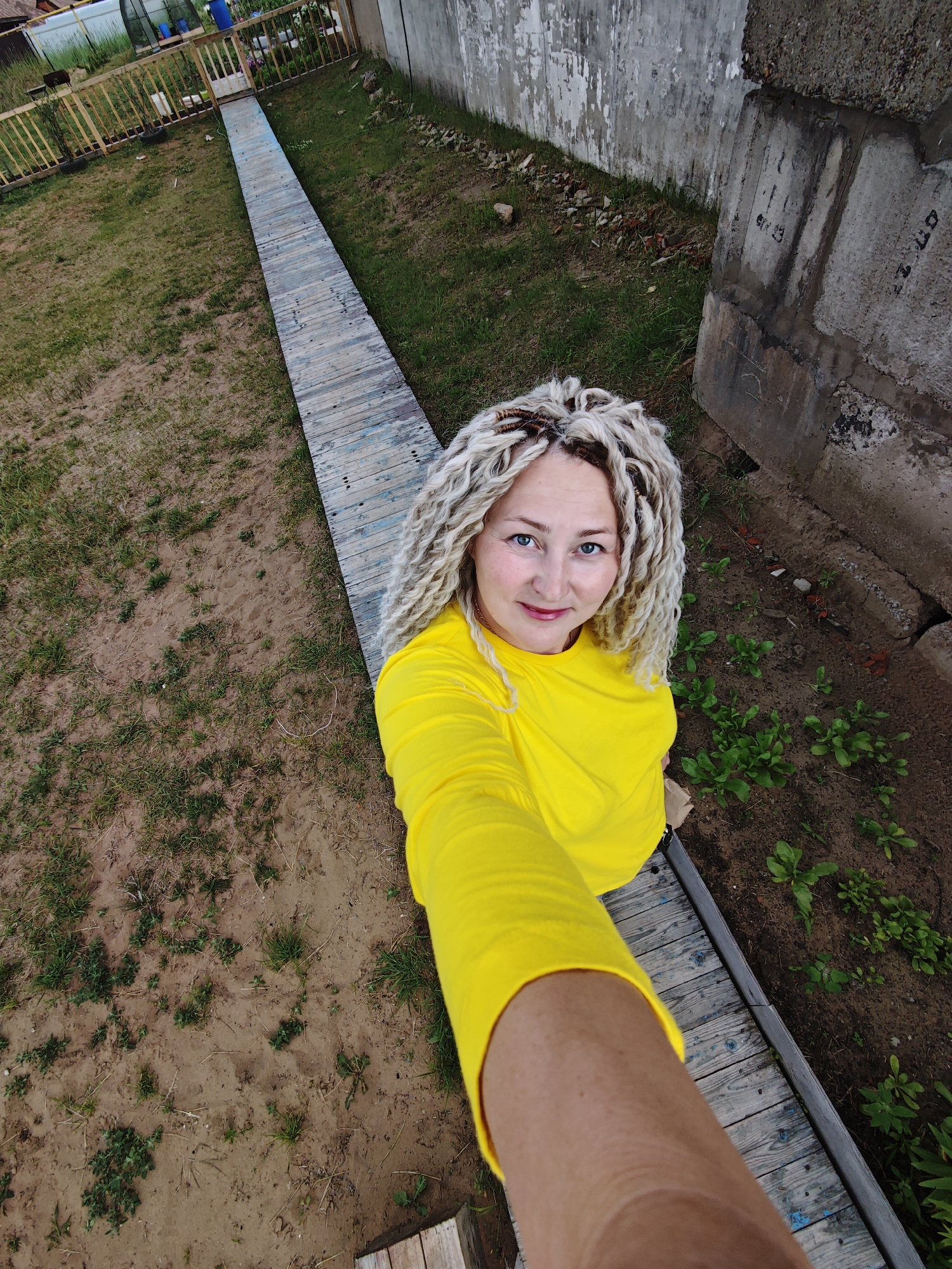 An adult Caucasian female with blonde braided hair is taking a selfie in an outdoor, rural setting. She is wearing a bright yellow long-sleeved shirt. She is standing on a wooden pathway that runs alongside a garden bed with small green plants. To her right is a concrete wall with some graffiti. The ground is a mix of soil and sparse grass. In the background to the left, a wooden fence and some structures suggest a backyard or dacha area. The overall environment appears to be a residential area outside of the city center.