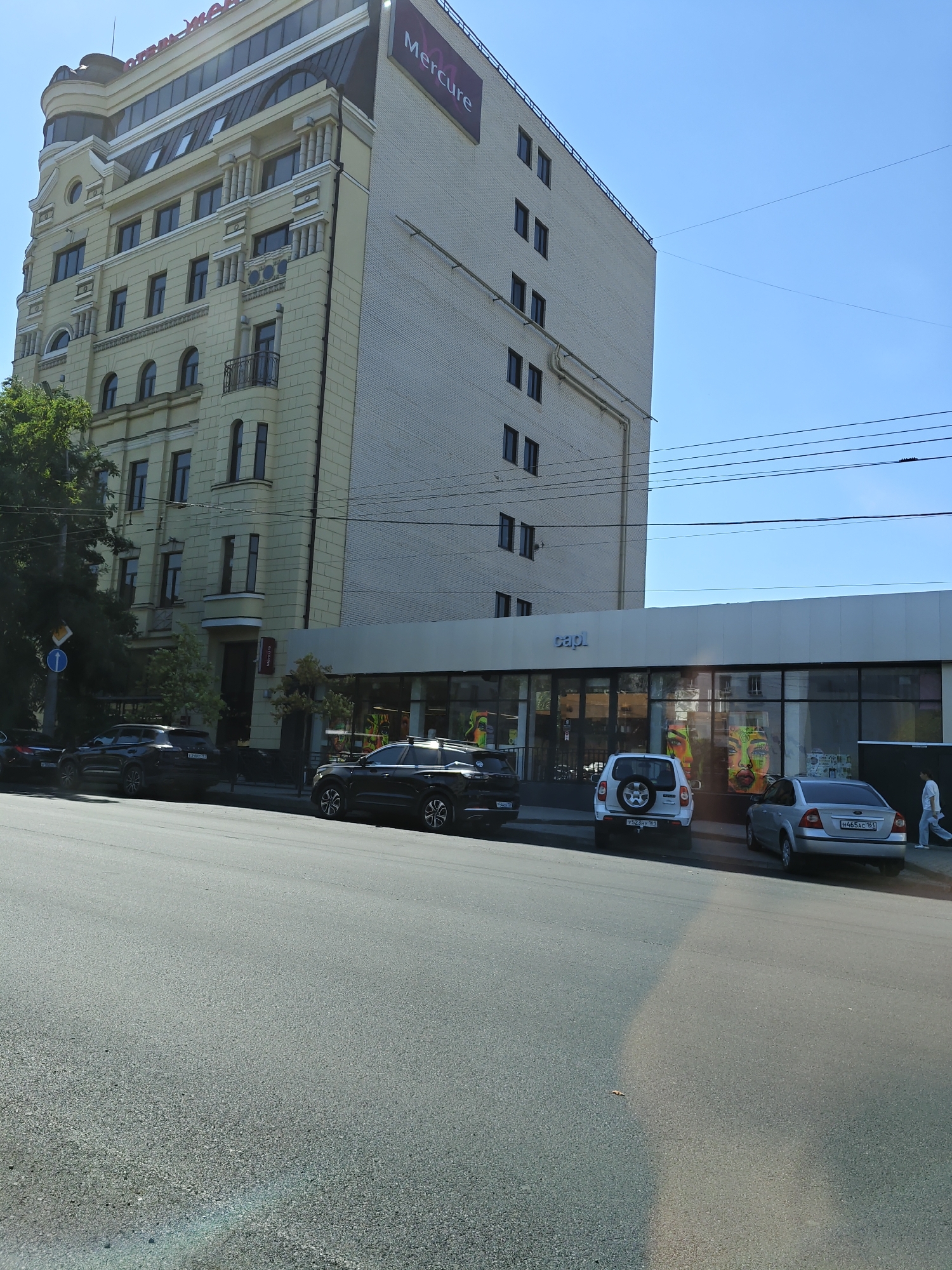 A three-story building with a yellow facade stands next to a taller, white brick building under a clear blue sky. The yellow building features architectural details like columns and rounded windows. The white building has a prominent "Mercure" sign and a series of evenly spaced dark windows. At the base of the white building is a commercial space with large glass windows displaying colorful artwork. Several cars are parked along the street in front of the commercial space, including two dark SUVs, a white SUV, and a silver sedan. A person in a white outfit is walking away from the camera on the sidewalk. A traffic sign is visible on the left side of the image. The street in the foreground is asphalt.
