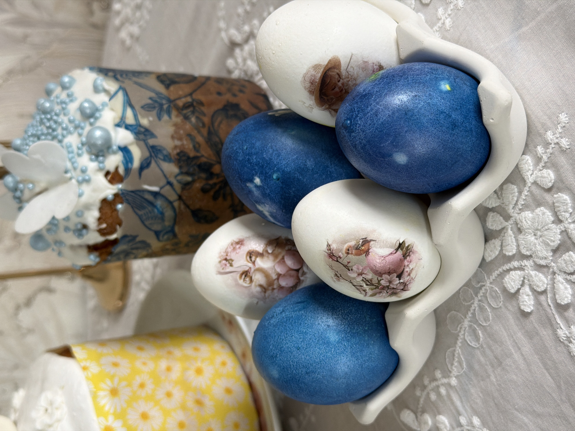 The image depicts a festive Easter still life. Three blue-dyed Easter eggs alternate with three white Easter eggs decorated with vintage-style decoupage, featuring images of birds and people. These eggs are held in a white ceramic egg holder. A partially visible Easter cake decorated with white frosting, blue sugar pearls, and white sugar flowers is in the background, to the left of the eggs. A yellow container with a daisy pattern can be seen at the base of the Easter cake. The composition rests on a white lace tablecloth. The location is likely in Reshetnikovo, Russia.
